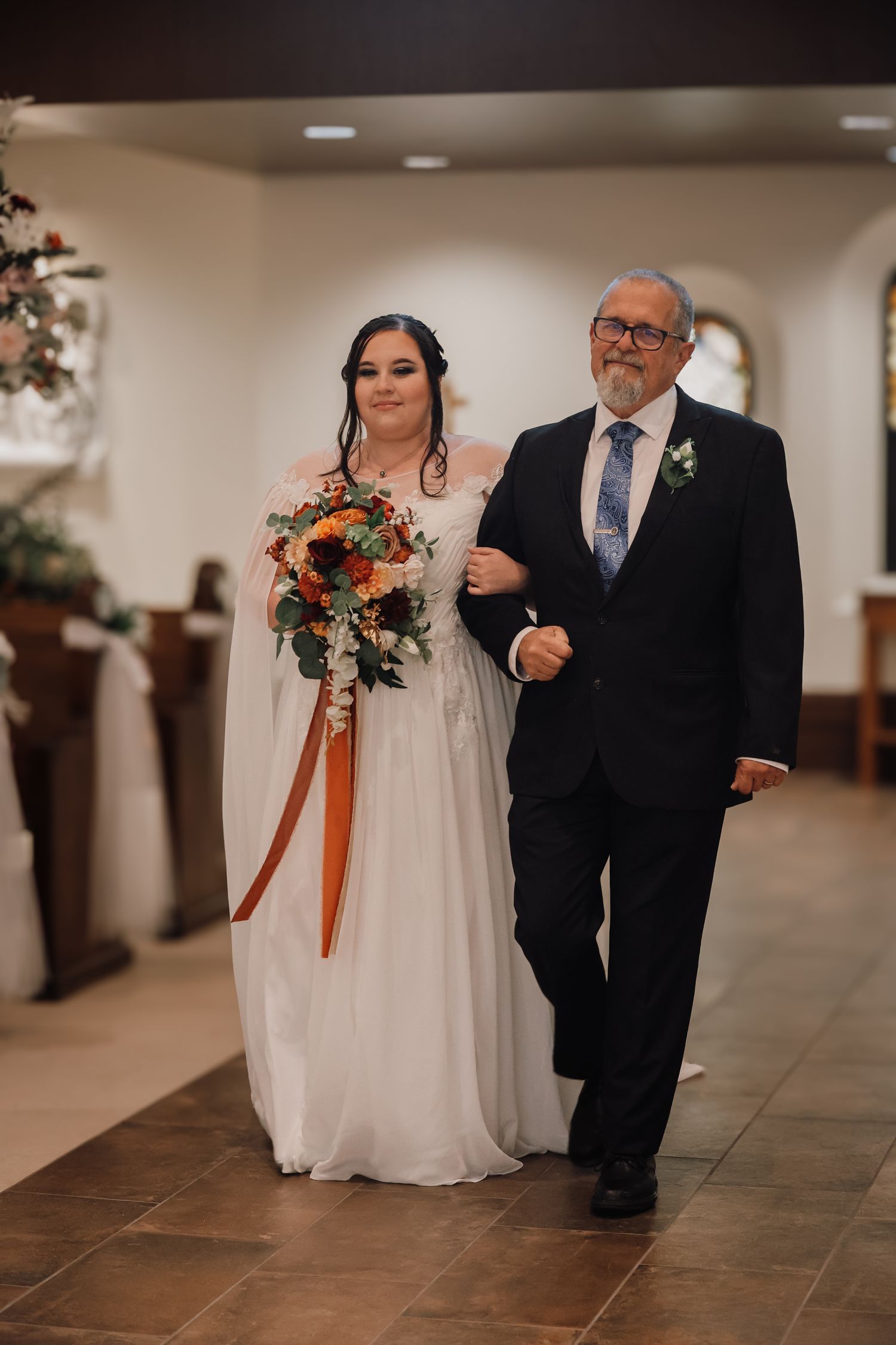 A bride in a white wedding dress walks down a church hallway with an escort in a dark suit.