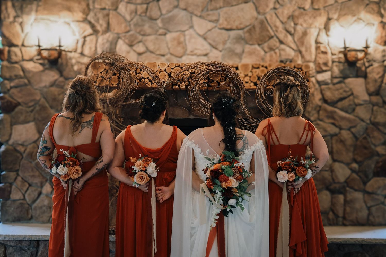A wedding party in rust-colored bridesmaid dresses stands against a stone wall holding autumn-toned bouquets.
