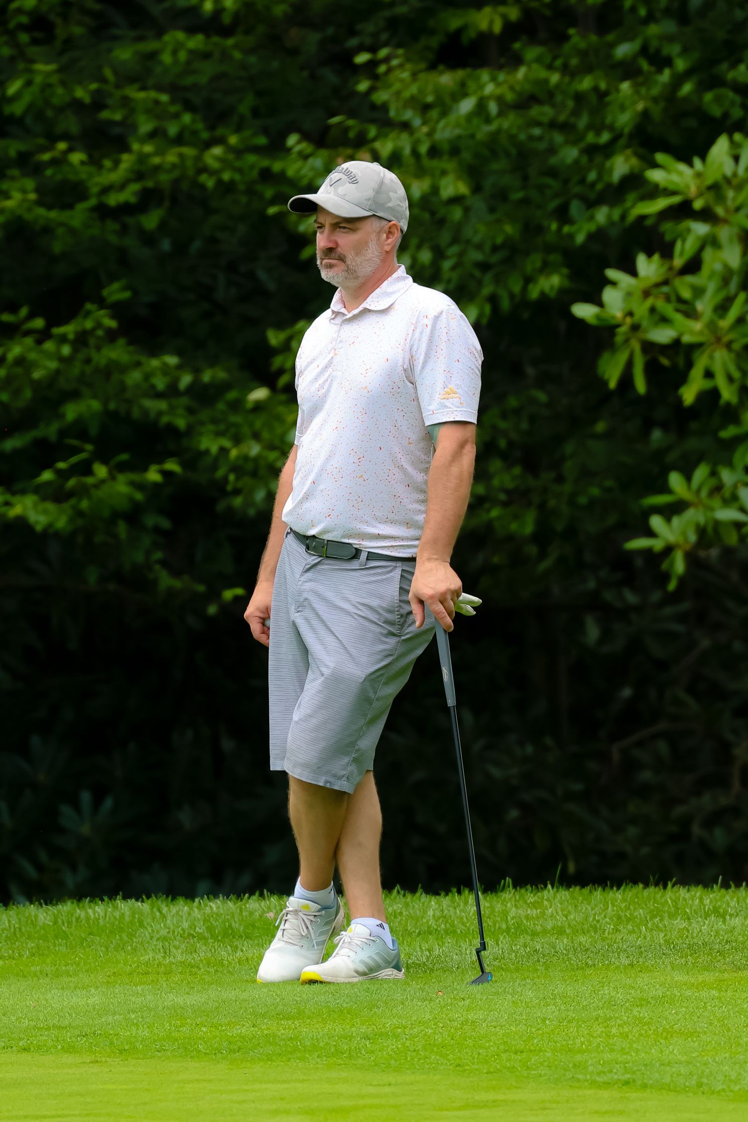Golfer in white shirt and gray shorts stands on lush green course preparing to putt.