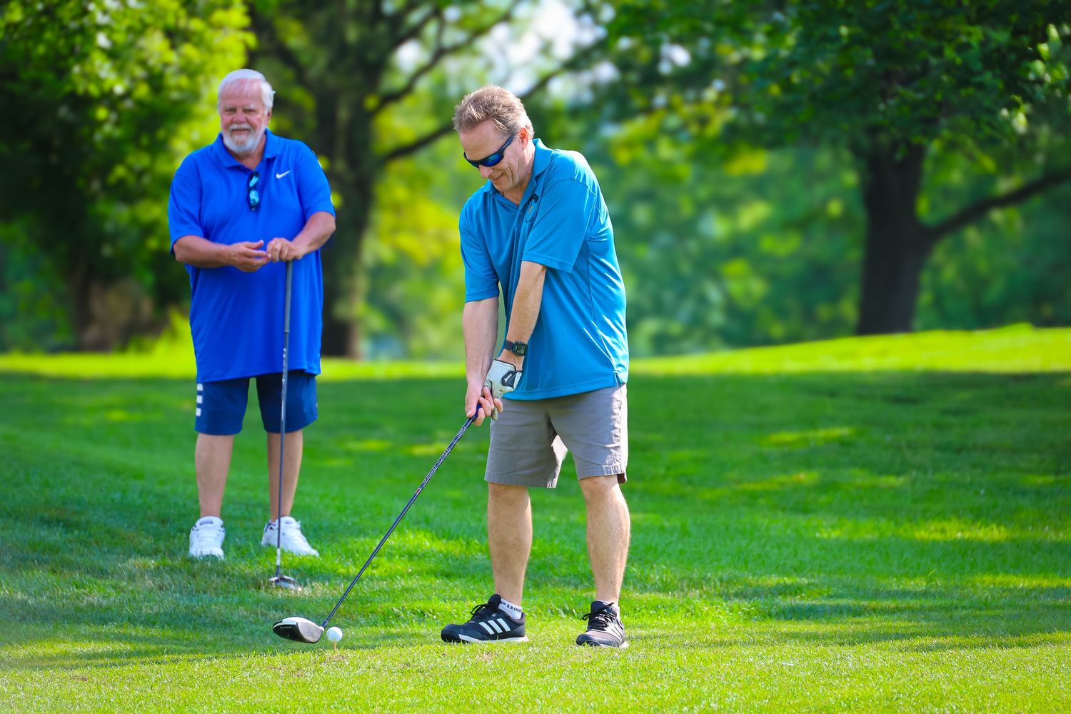 Two golfers on a lush green course, one preparing to tee off while the other watches on a sunny day.