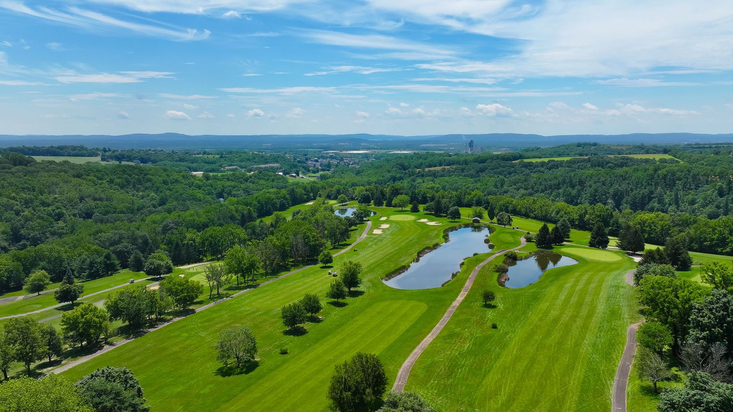 Panoramic aerial view of a lush green golf course with fairways, trees, and water hazards under bright blue skies.
