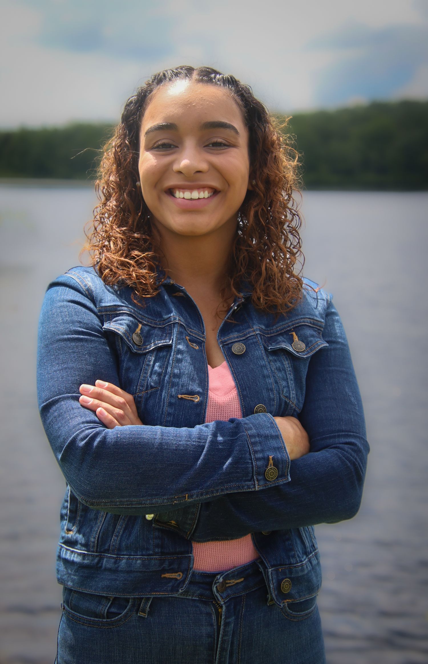 Smiling woman with curly hair wearing denim jacket stands with arms crossed by a lake on cloudy day.
