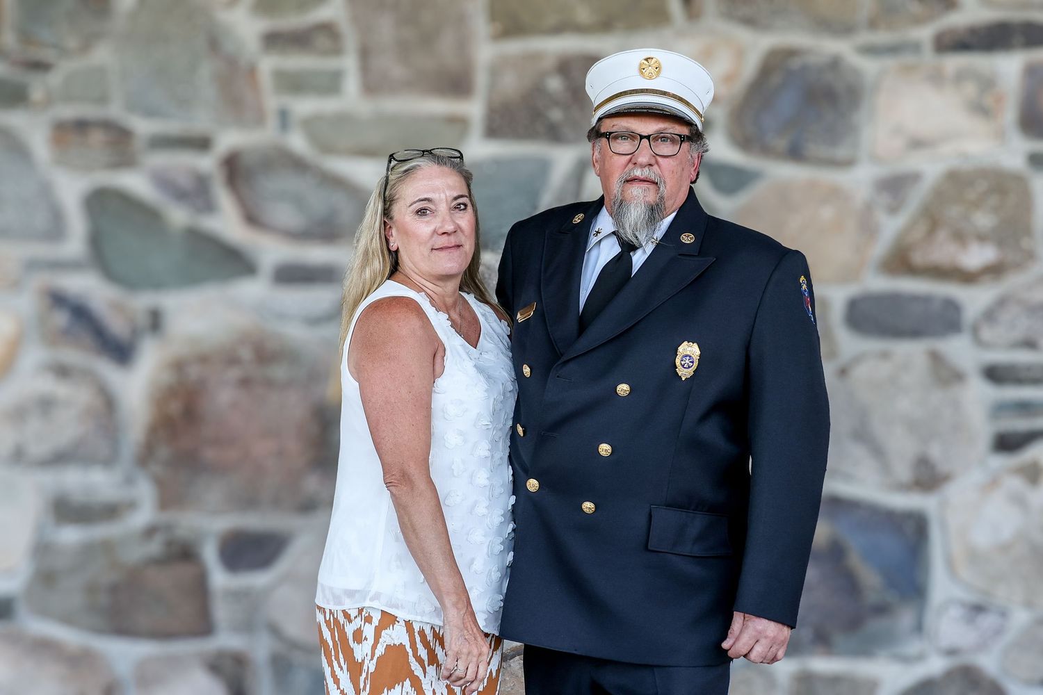 A couple poses together against a stone wall, with one wearing a white dress and the other in a formal navy uniform.