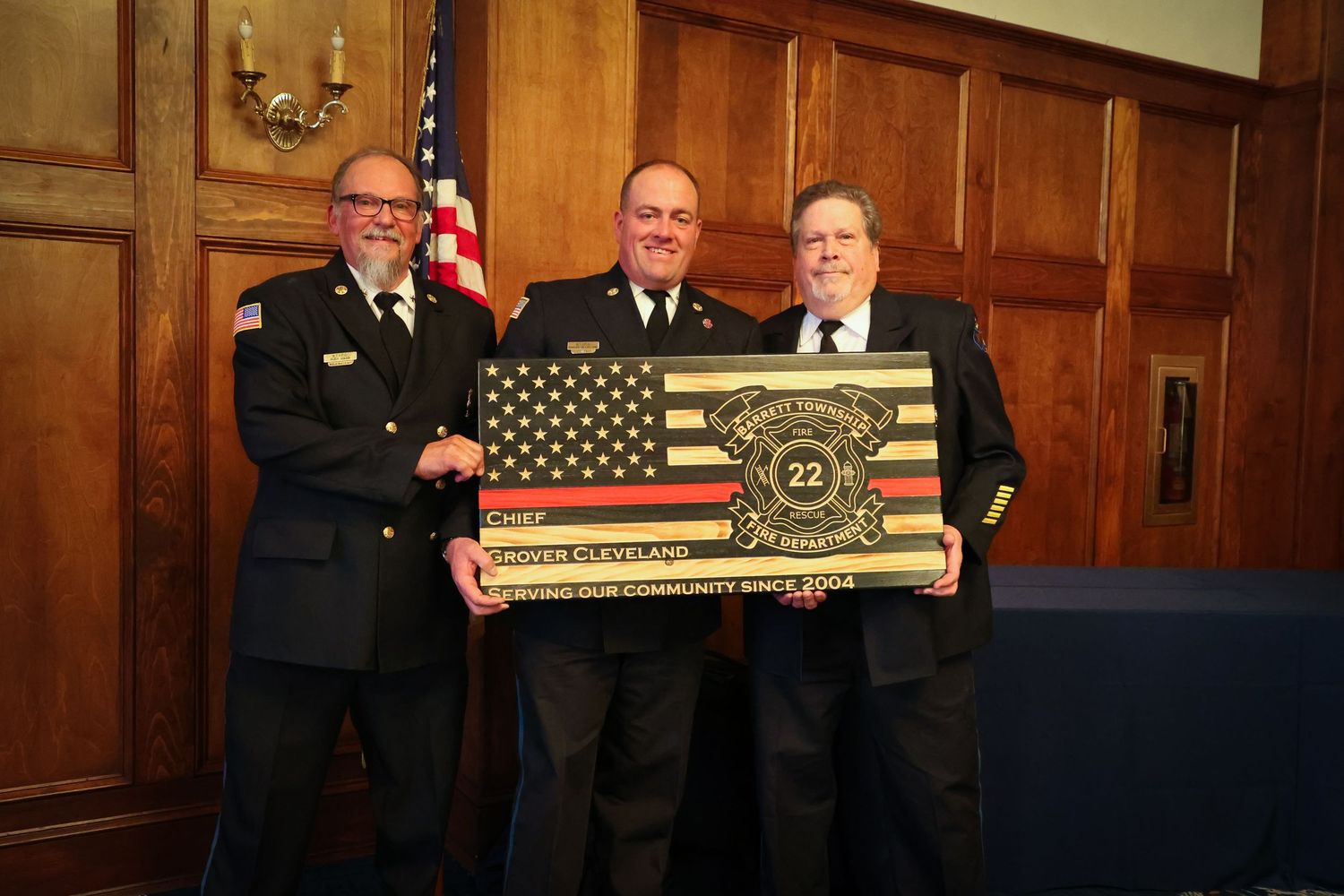 Military personnel stand in a formal ceremony holding ceremonial oars with American flags in a wood-paneled room.