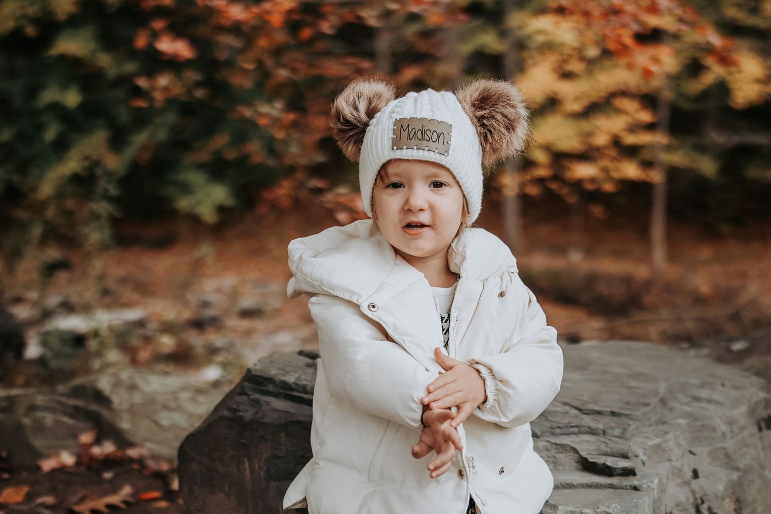 A small figure in a white winter coat and hat explores a rocky area surrounded by autumn foliage.
