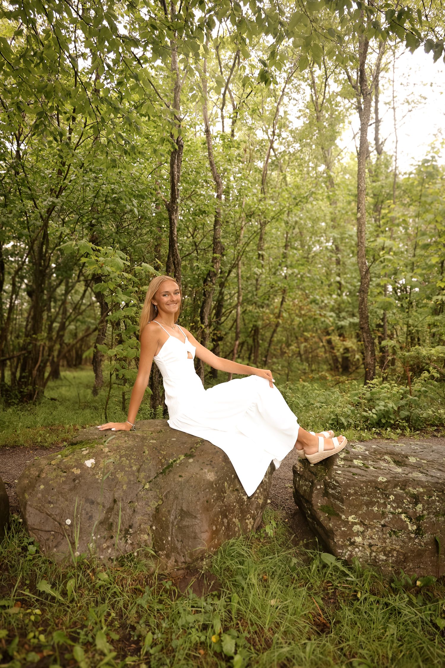 A woman in a white dress reclines gracefully on a large rock in a wooded forest setting.