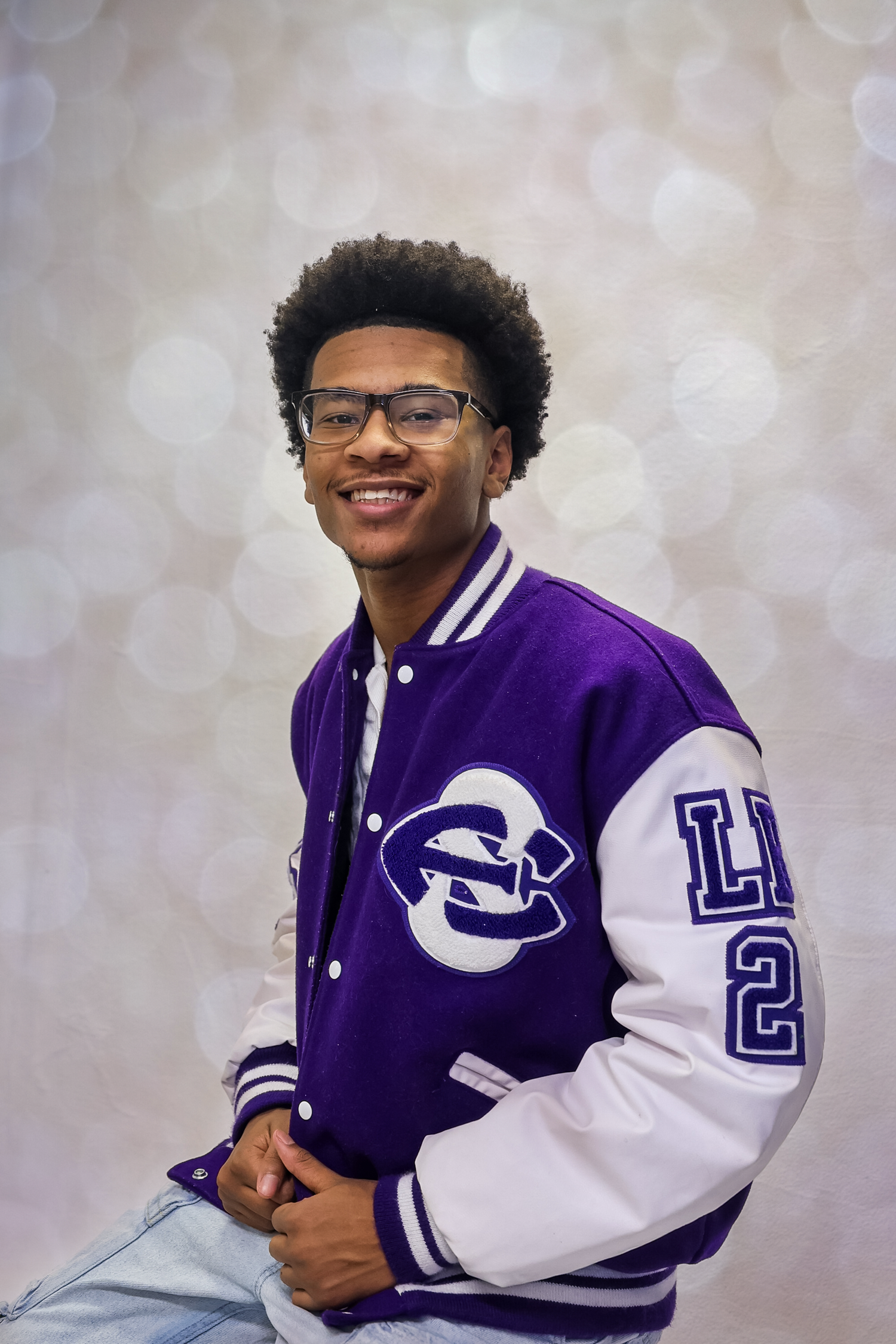 A student wearing a purple and white varsity letterman jacket with glasses poses against a light background.