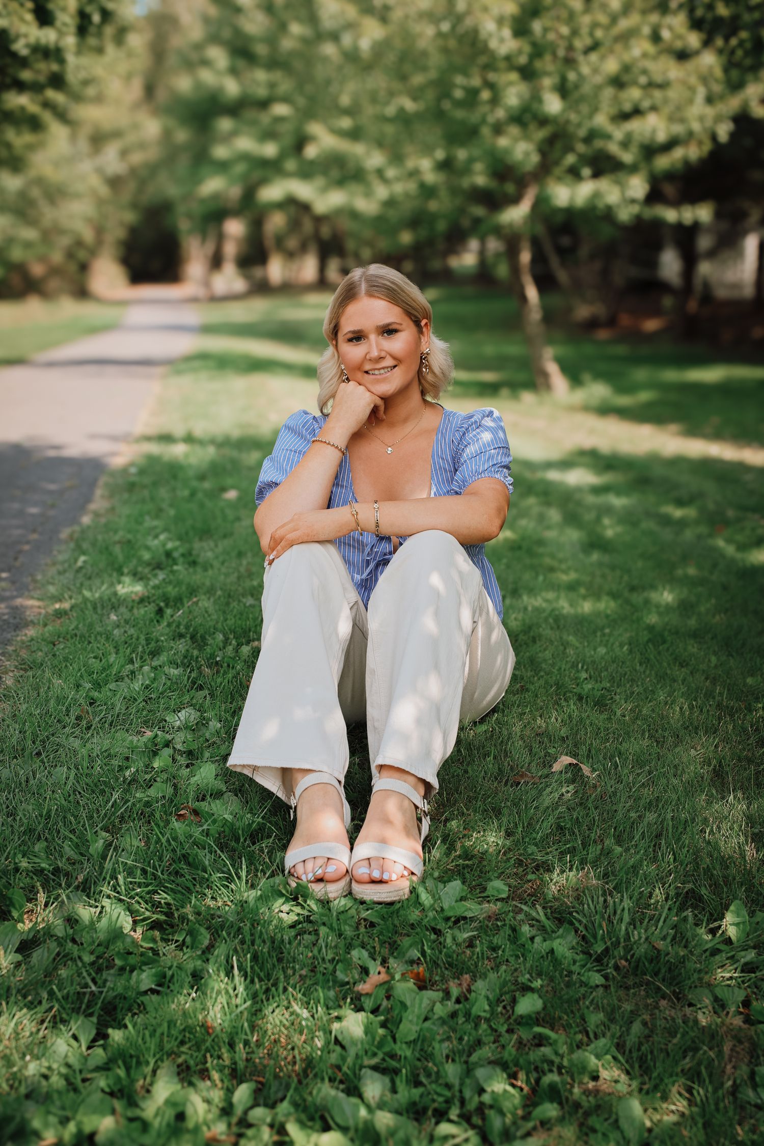 A person in a blue top and white pants sits on lush grass in a sunny park setting.