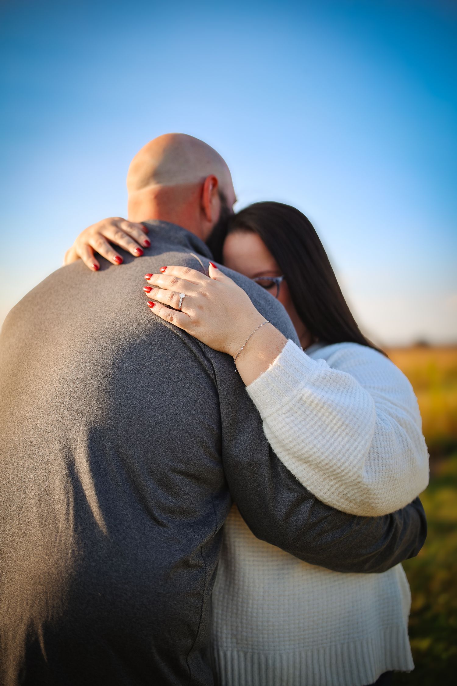 A romantic couple embraces in a field at sunset, with one wearing a white sweater and the other in grey.