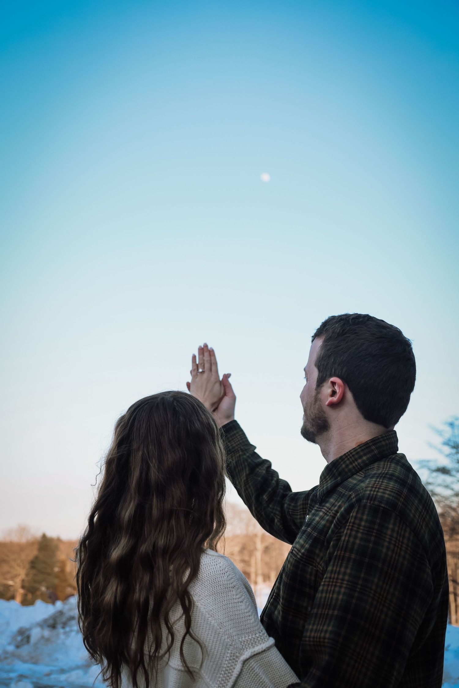 A couple in silhouette points at the moon in a bright blue sky during daytime.