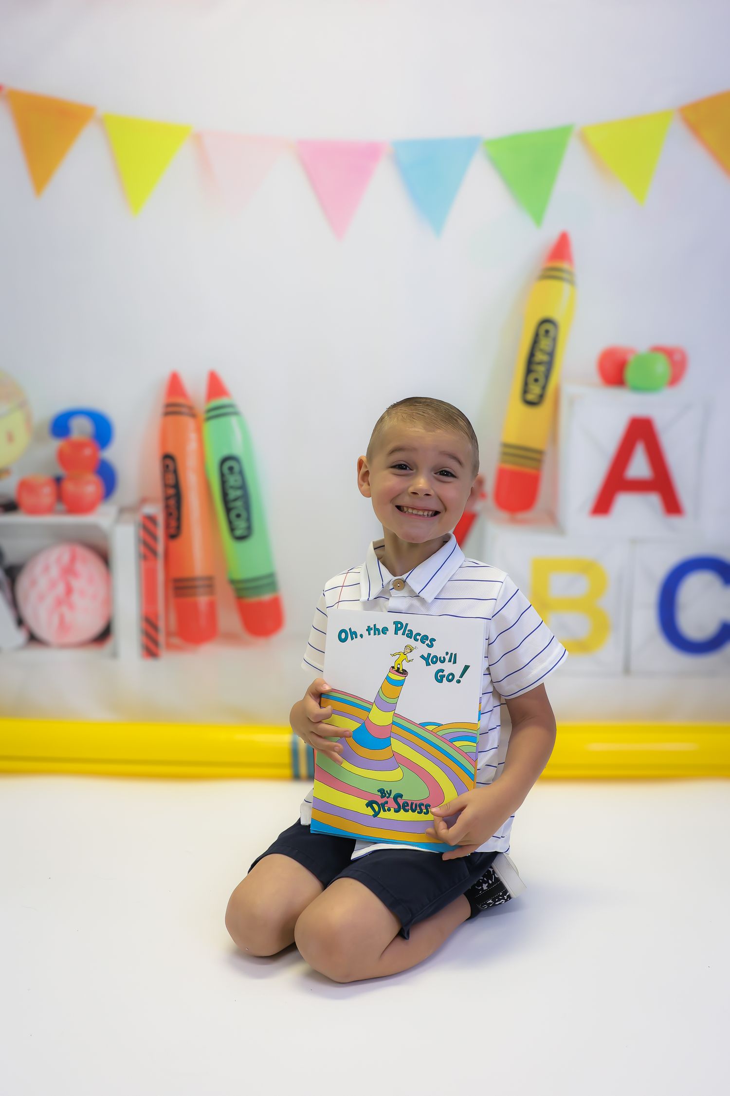 Smiling boy kneeling holding Dr. Seuss Oh the Places Youll Go book with colorful crayon backdrop.