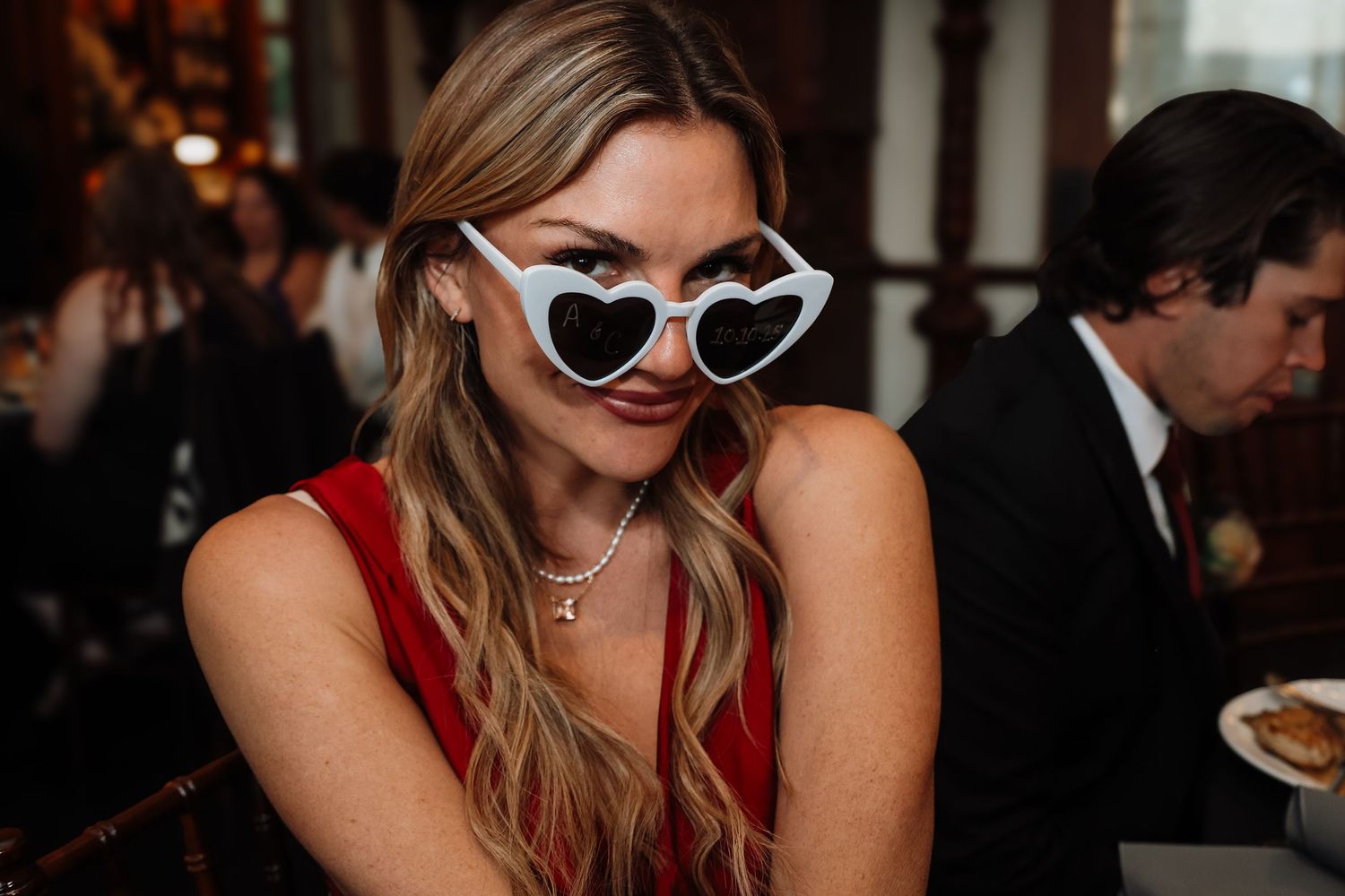 A person in a red dress and heart-shaped sunglasses smiles while dining at a restaurant.