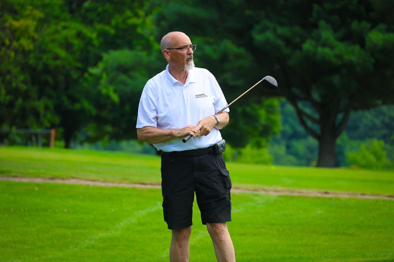A golfer in a white shirt takes a swing on a lush green golf course during a summer day.