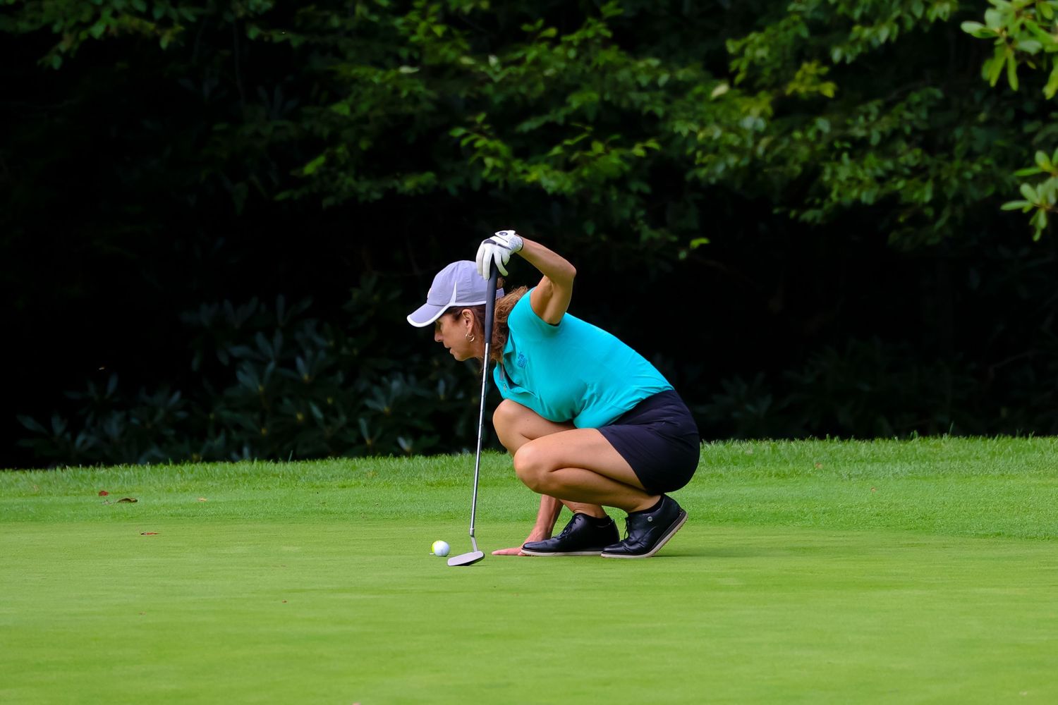 Golfer in teal shirt and navy shorts crouches to read the green on a sunny golf course.