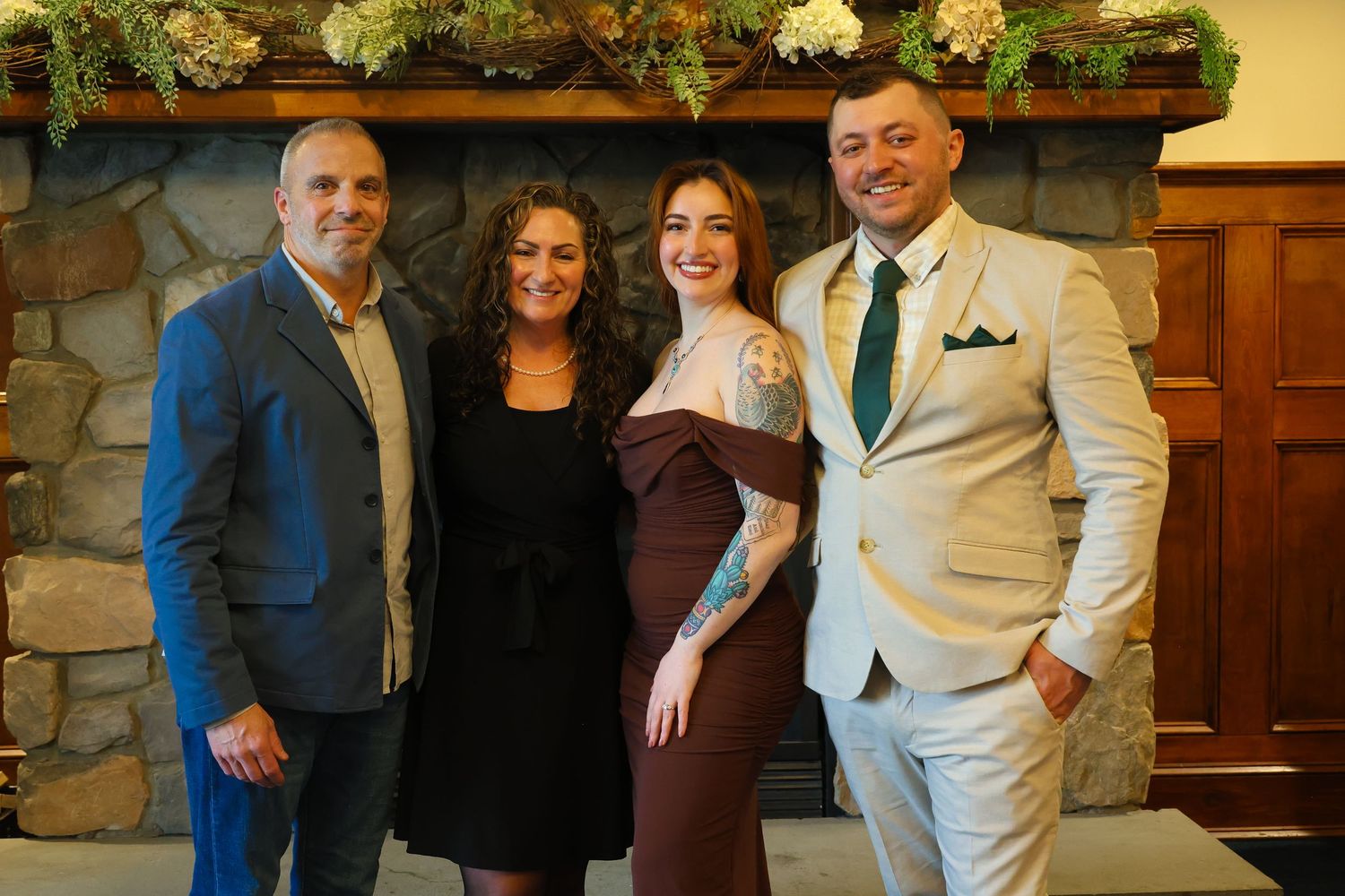 People in formal attire gather together for photos at an indoor event with wood paneling and stone walls in the background.