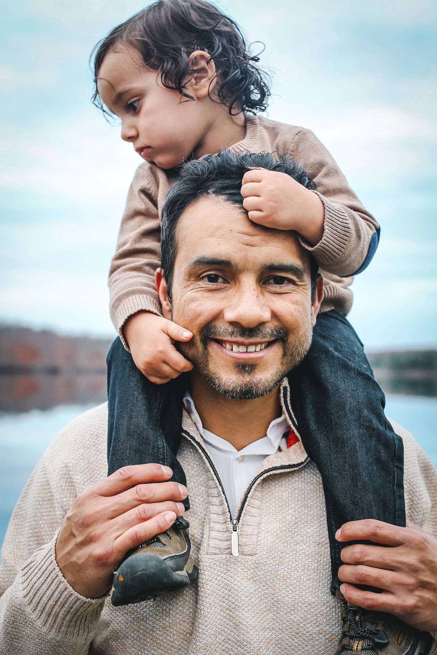 A parent carries a young child on their shoulders while wearing a beige sweater against a blue sky background.