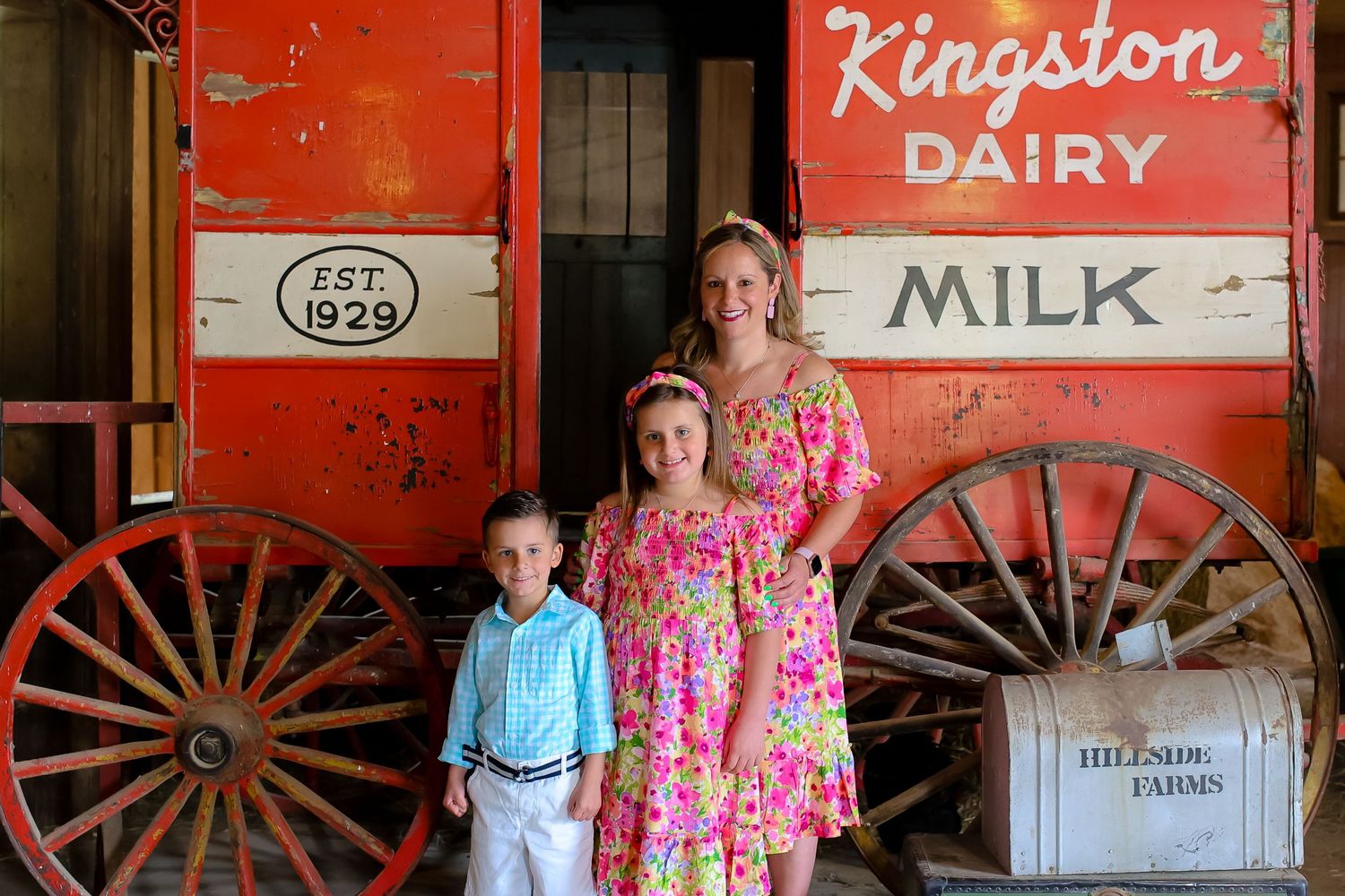 A vintage red barn wall displays Kingston Dairy signage alongside rustic wooden wagon wheels and milk advertising.