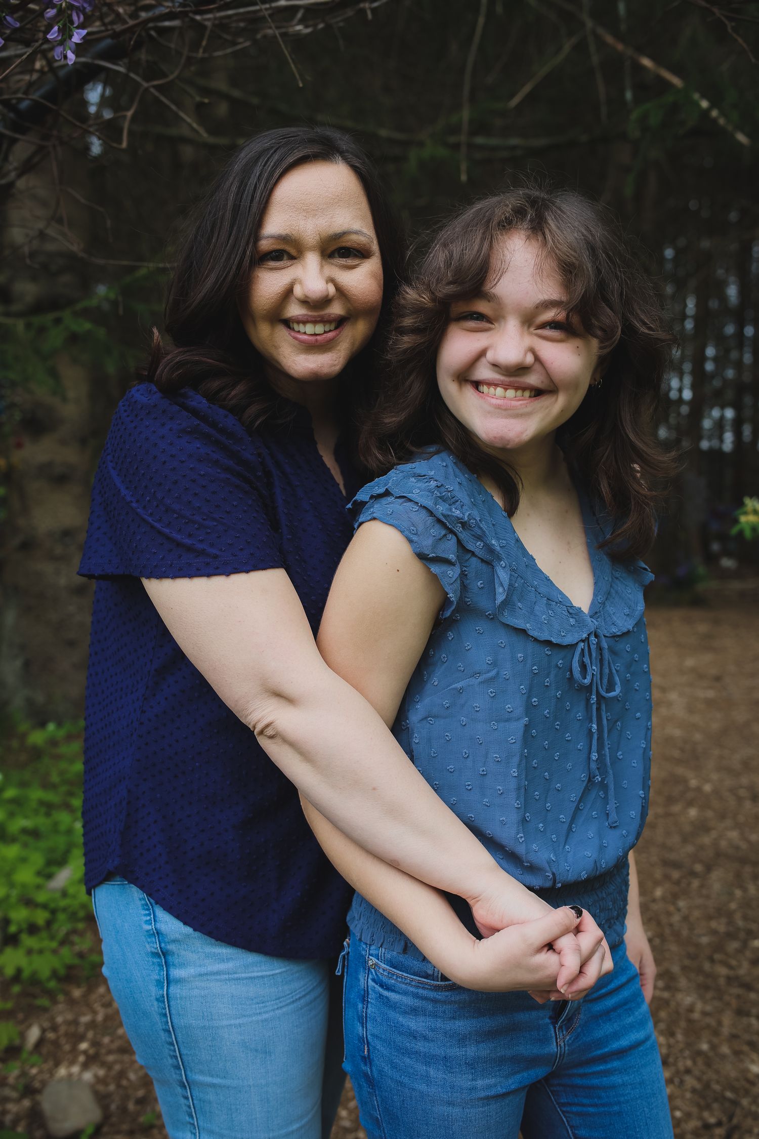 Two people wearing blue and navy tops embrace and smile while standing in a wooded outdoor setting.