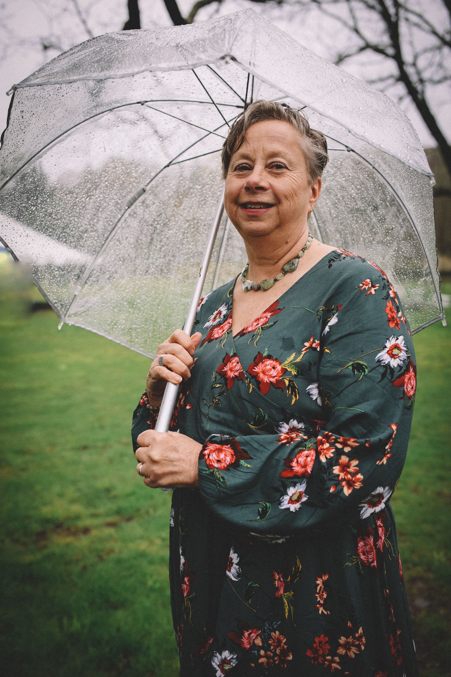 A person in a floral green dress smiles while holding a clear umbrella in a grassy outdoor setting during spring.