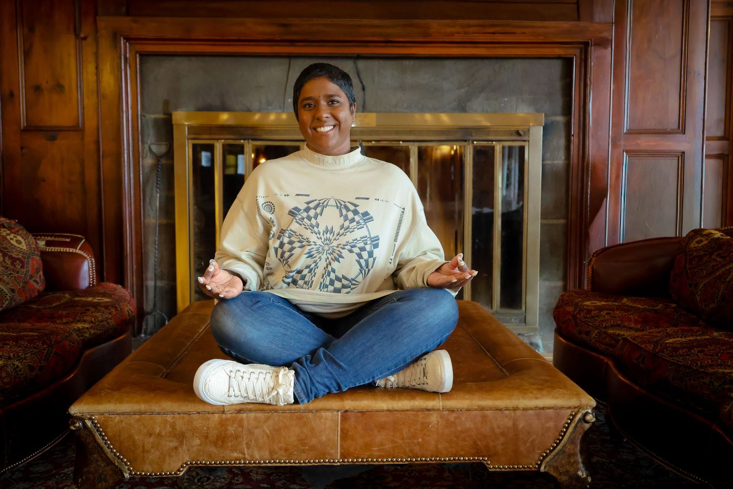 A person sits cross-legged on a vintage brown leather couch in a dimly lit room.