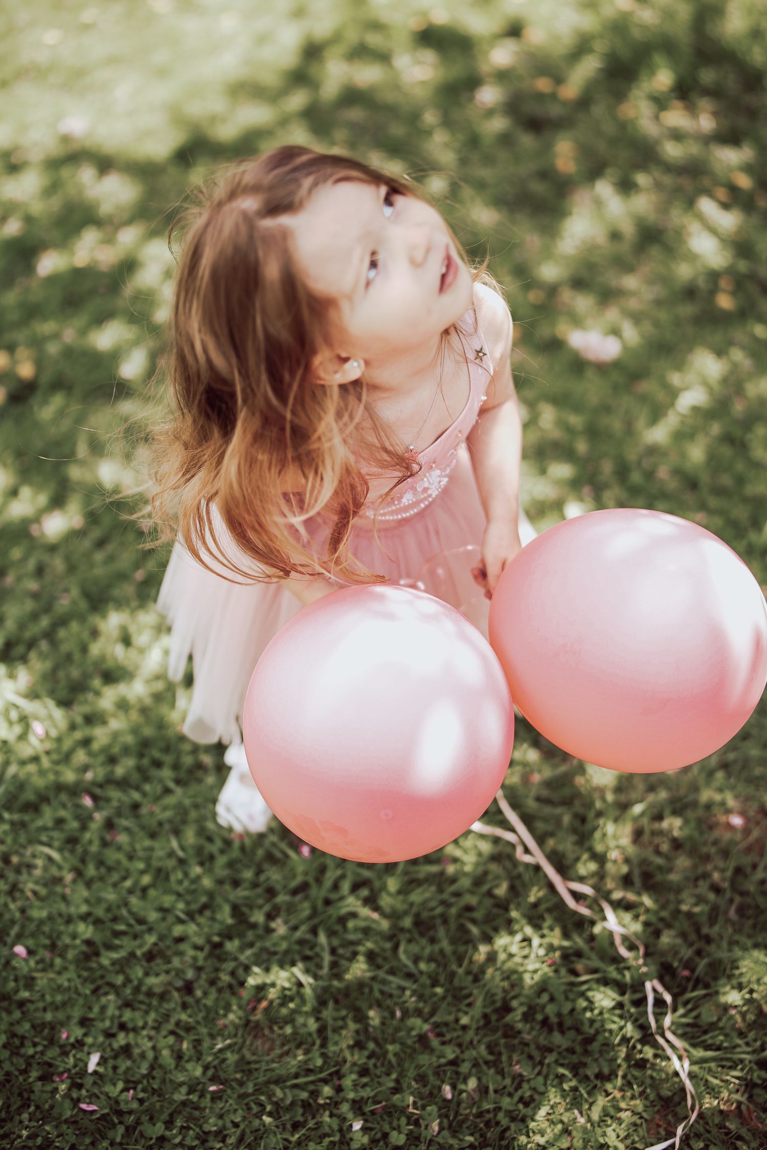 A child in a pink dress plays with pink balloons on grass in a whimsical outdoor photo series.