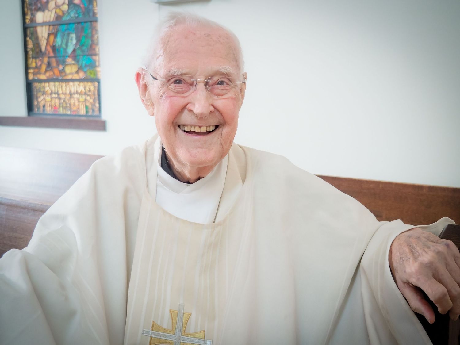 A priest in white robes smiles warmly while performing religious duties at the altar during a church service.