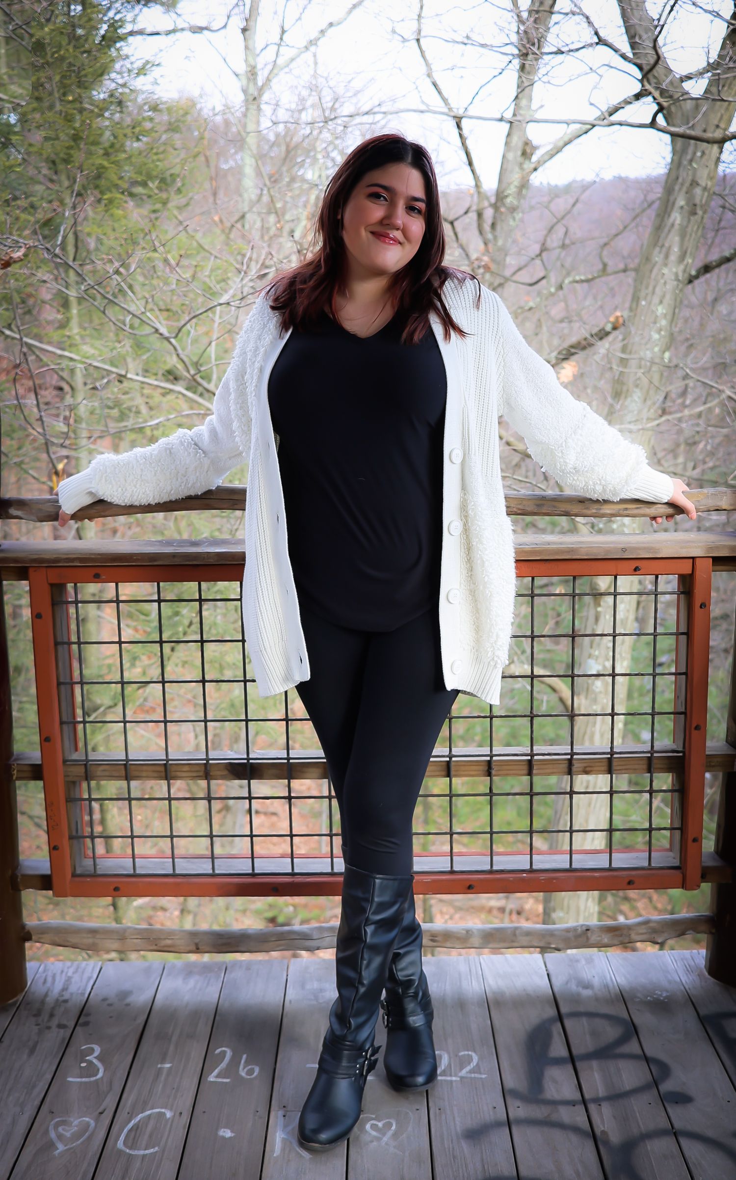 Someone in a casual black and white outfit stands on a wooden deck overlooking a scenic forest view in autumn.