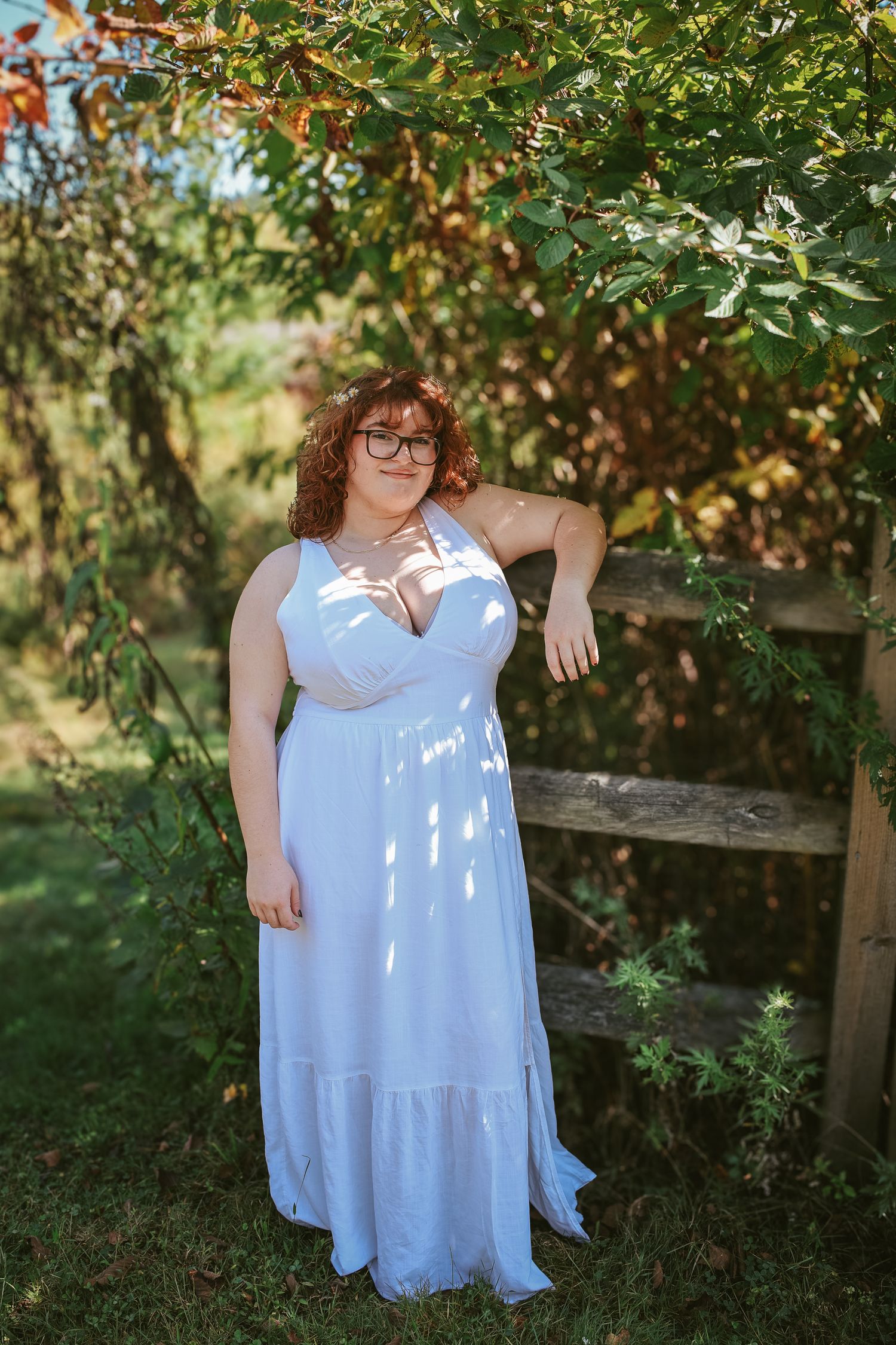 A person in a flowing white dress poses gracefully in a garden setting with dappled sunlight filtering through trees.