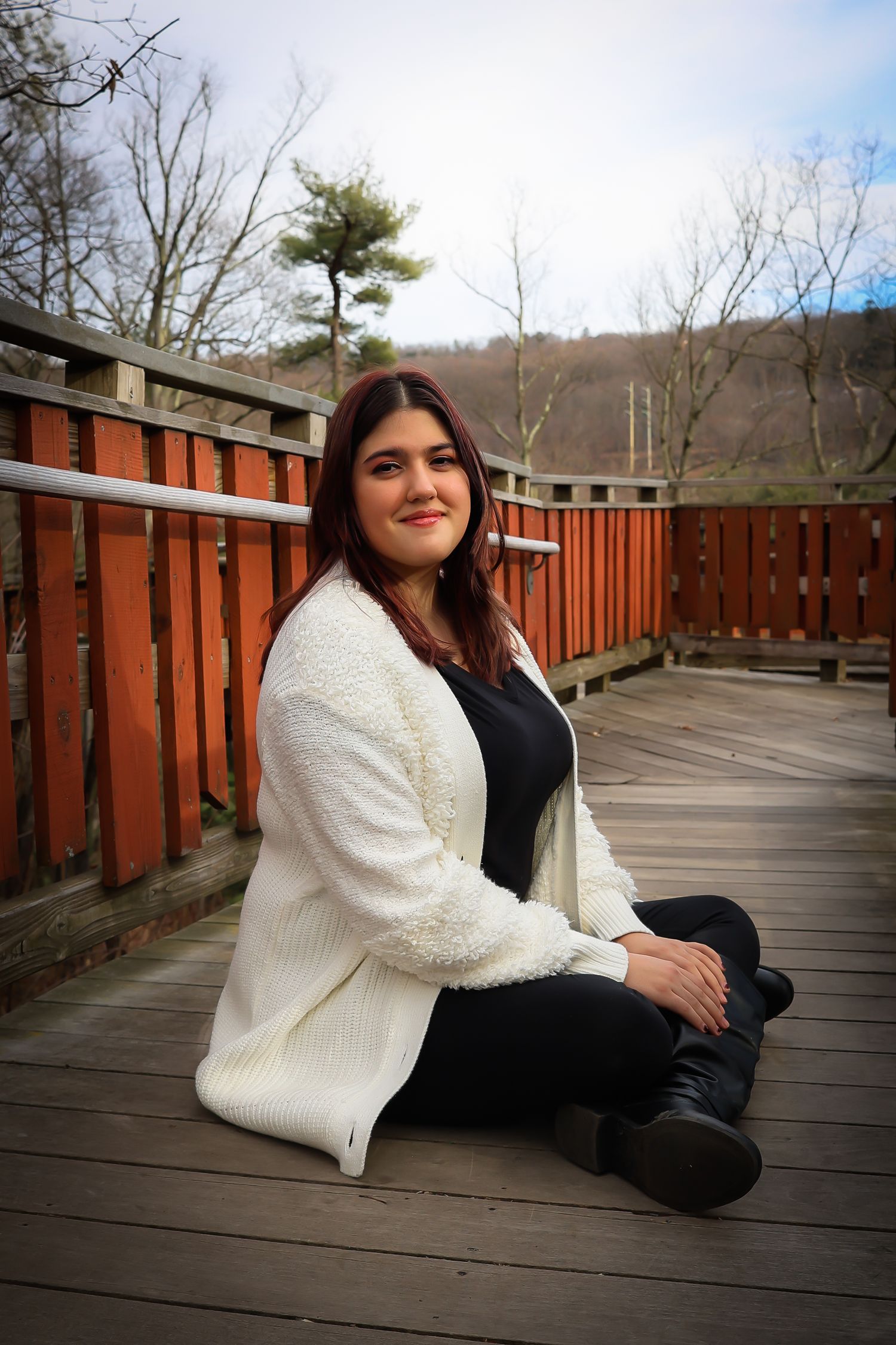 Person in a white fuzzy jacket and black outfit poses on a wooden deck with trees in the background.
