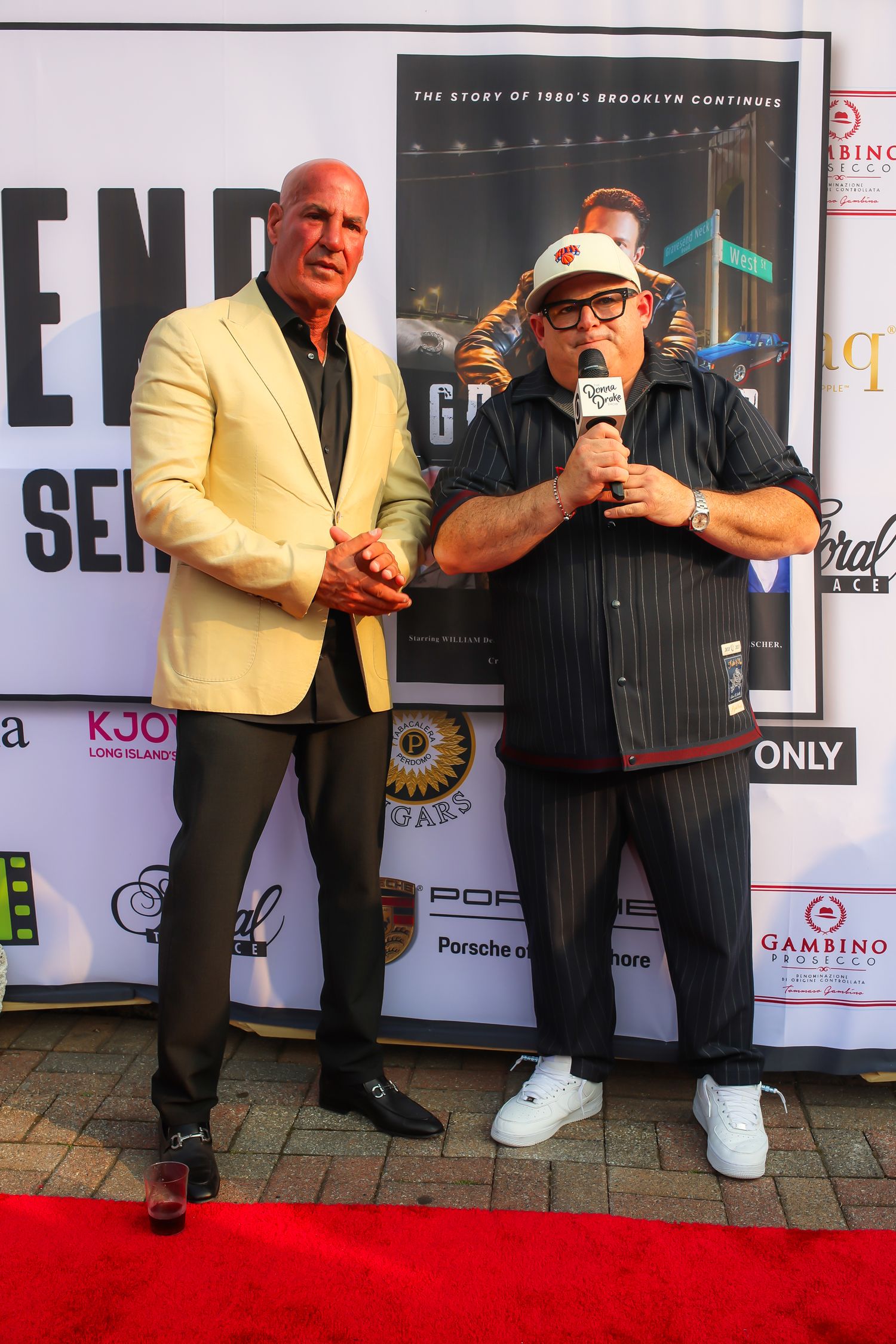 A person in a yellow blazer and dark pants poses with another person in black clothing on a red carpet.