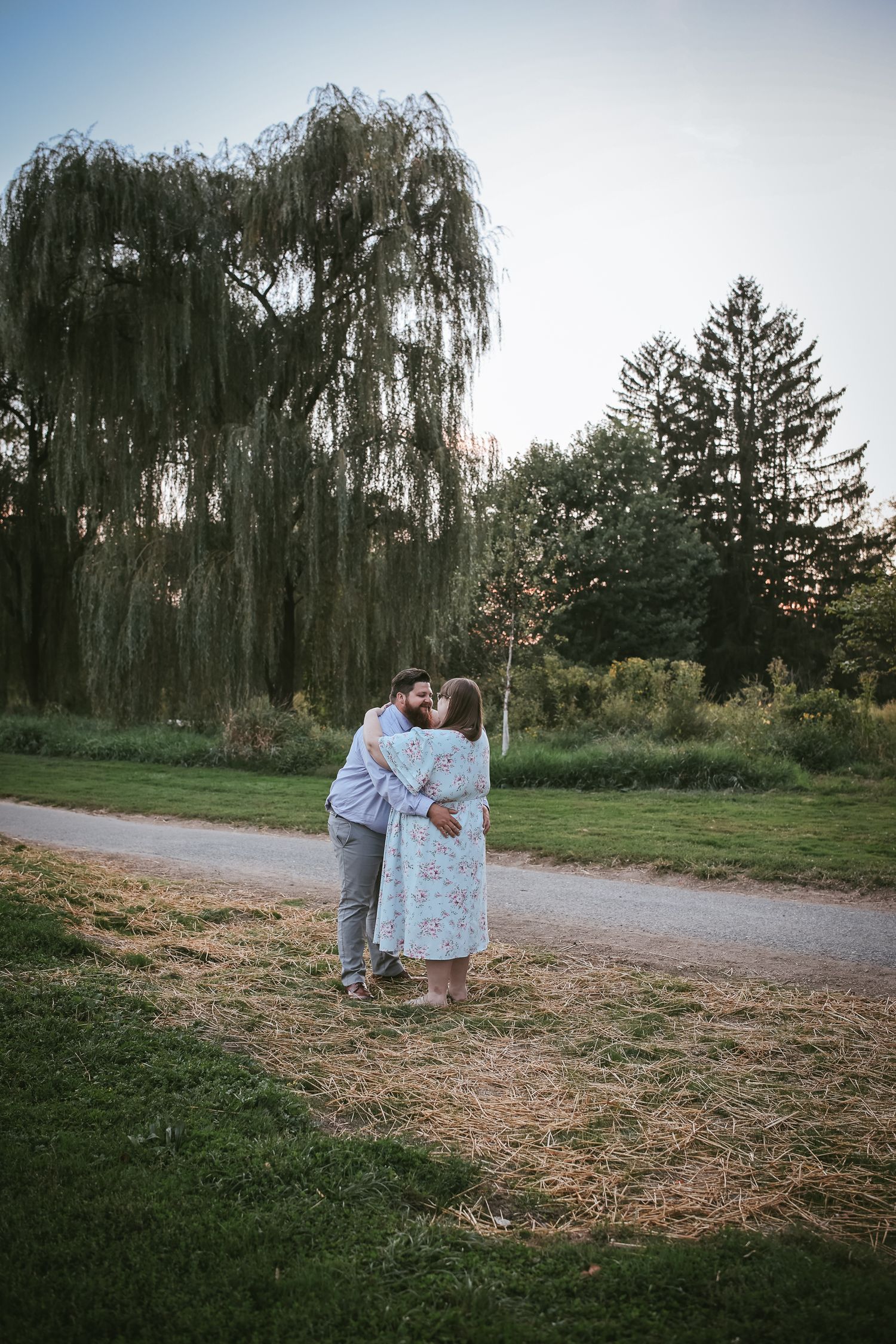 A couple shares a romantic moment under a weeping willow tree at dusk in a park setting.