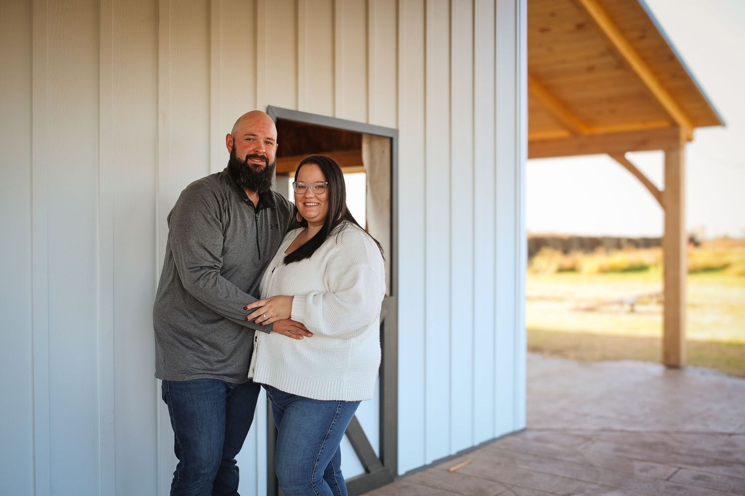 A couple stands together and embraces outside a white barn building on a sunny day.