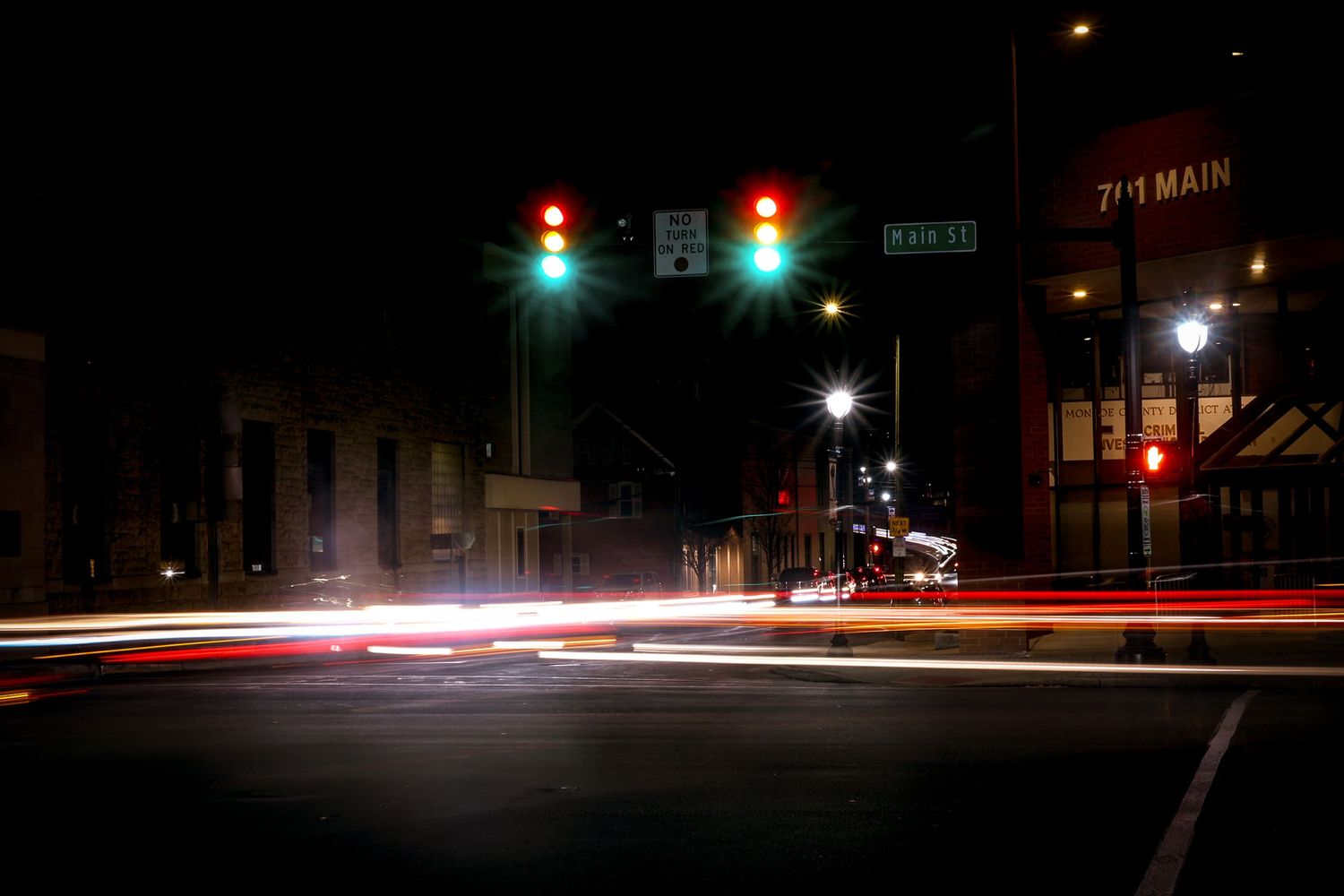 Long exposure night photography captures light trails at a city intersection with traffic signals and street lights.