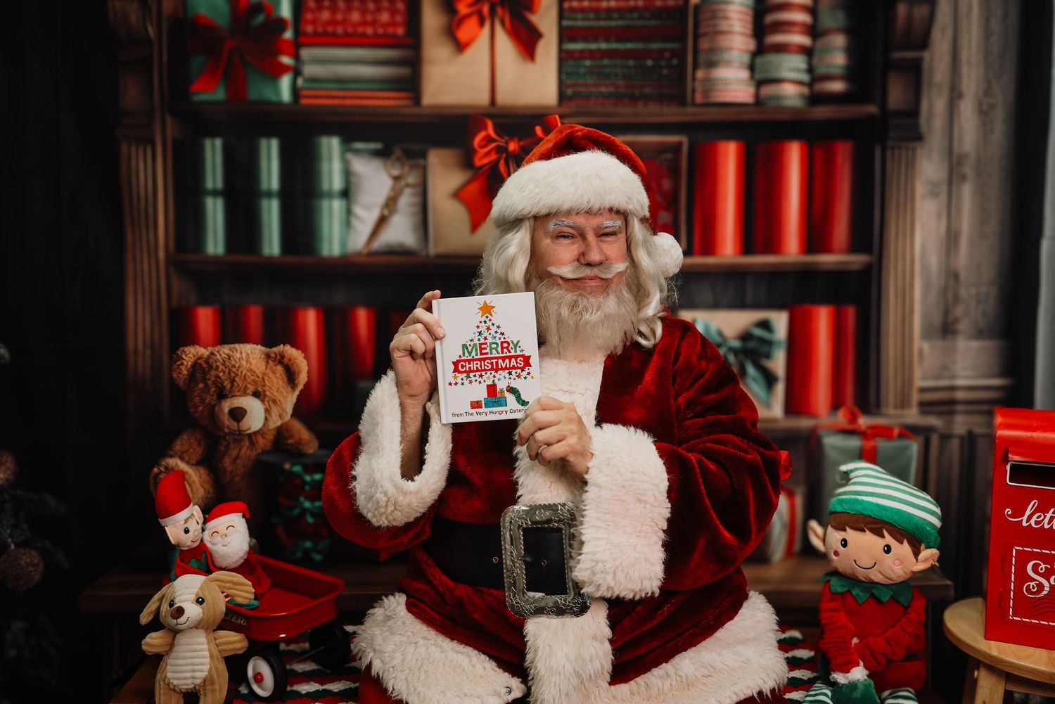 Santa Claus sits in a cozy holiday setting with red and white decor while taking a selfie on a smartphone.