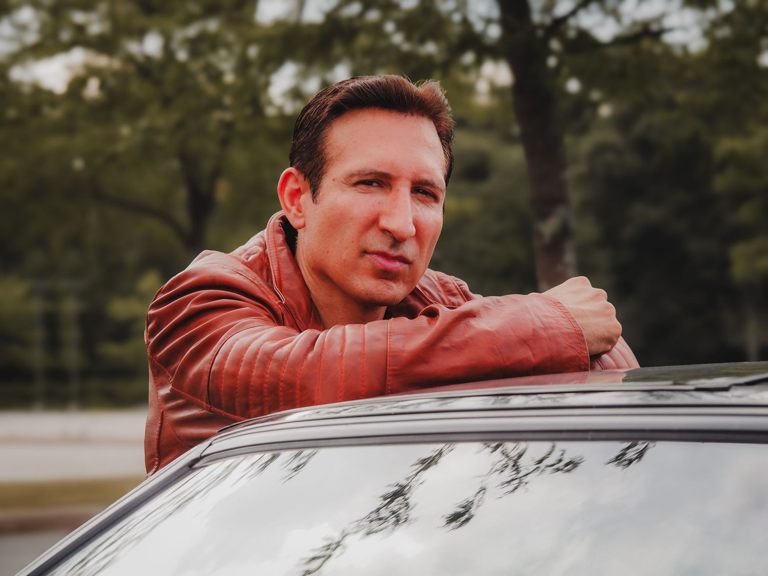 A person in a brown leather jacket leans against a car while posing for a series of outdoor photos.