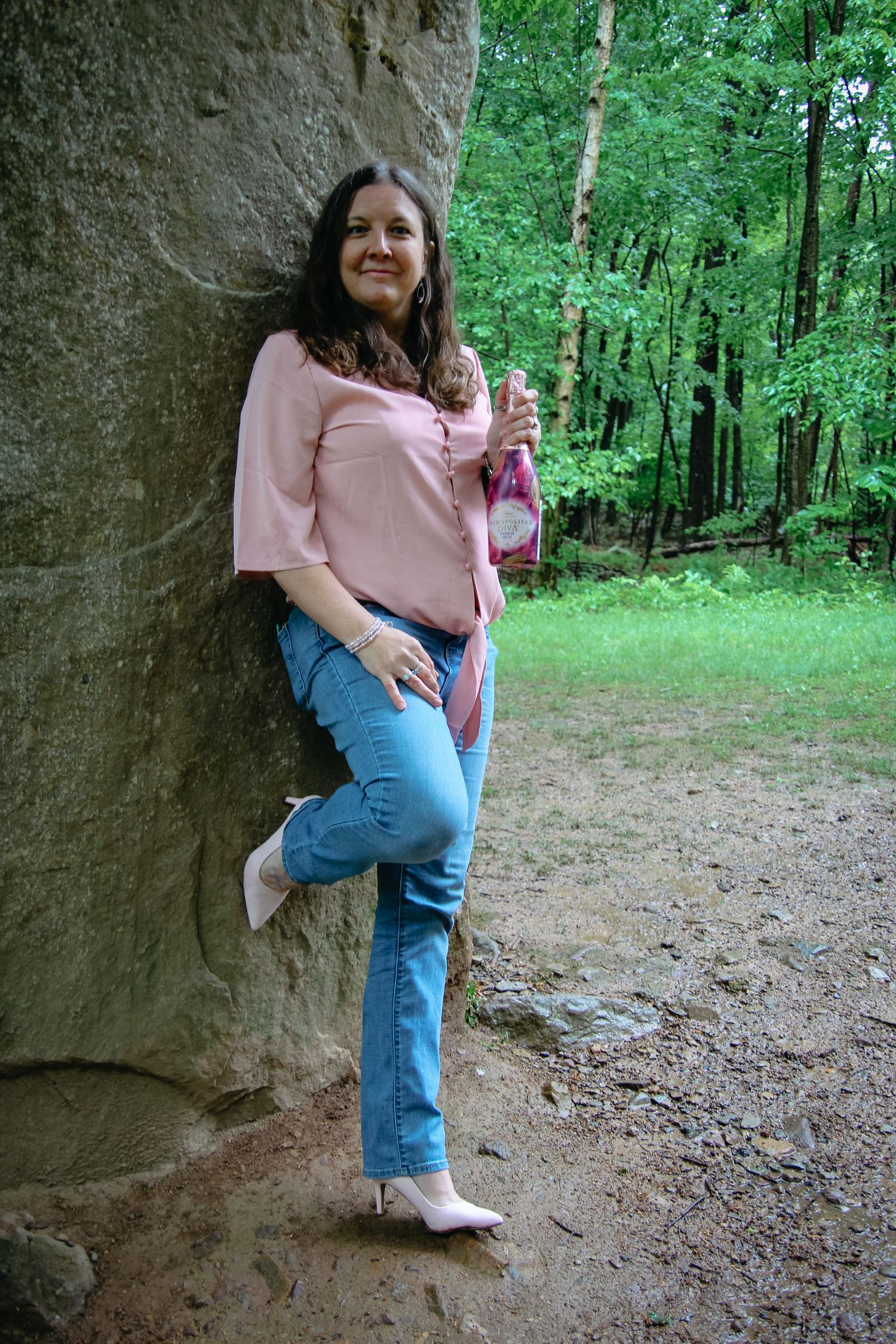 Someone in a pink top and jeans poses casually against a rock wall with lush green forest in the background.
