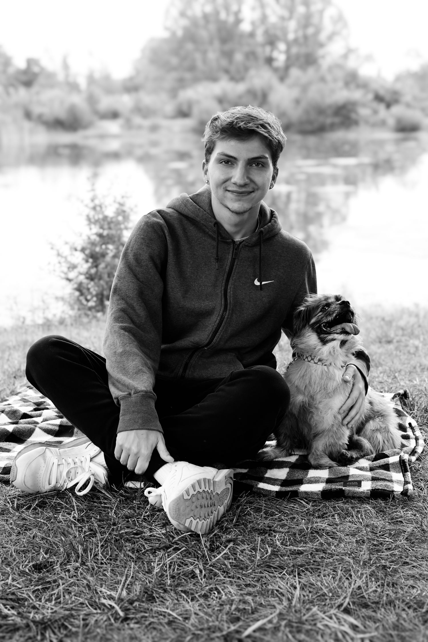 Black and white photo of someone in a Nike hoodie sitting on a blanket by a lake with their dog.