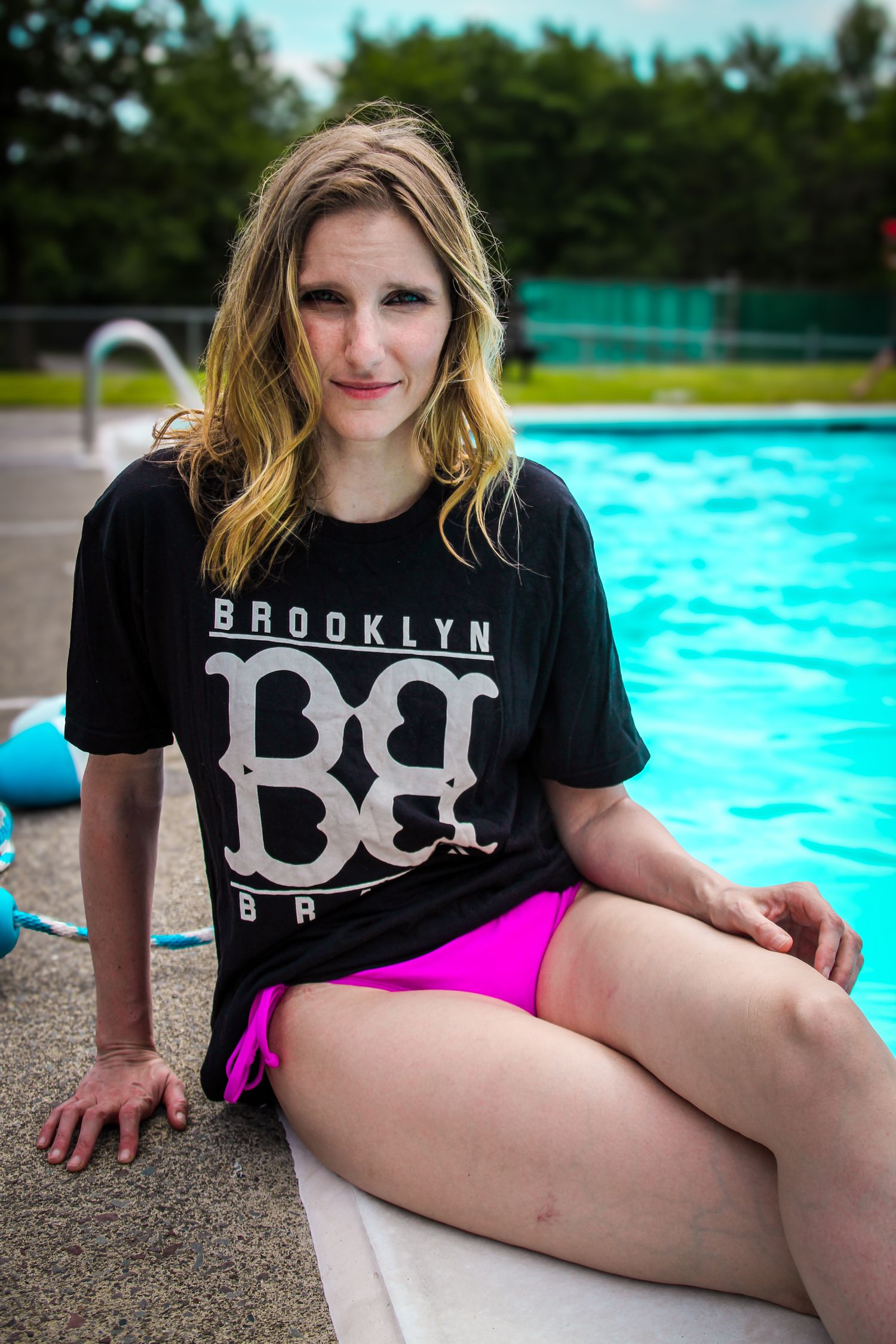 Someone wearing a black Brooklyn t-shirt and pink shorts sits at the edge of a bright blue swimming pool on a summer day.