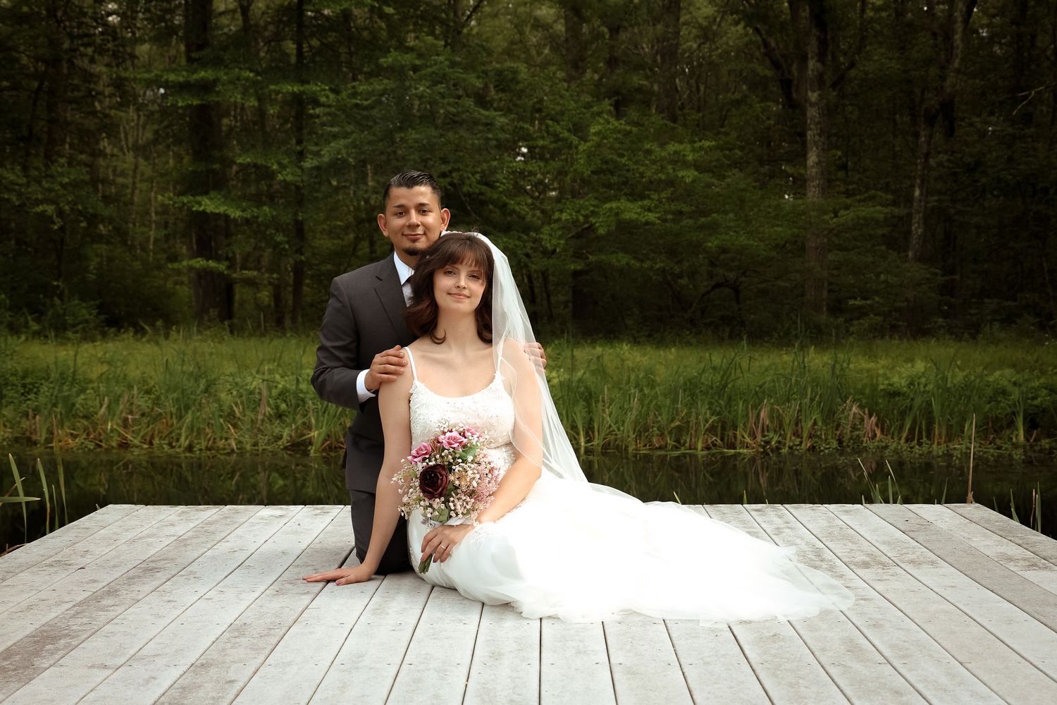 A bride and groom pose for romantic wedding photos on a wooden dock surrounded by lush greenery and trees.