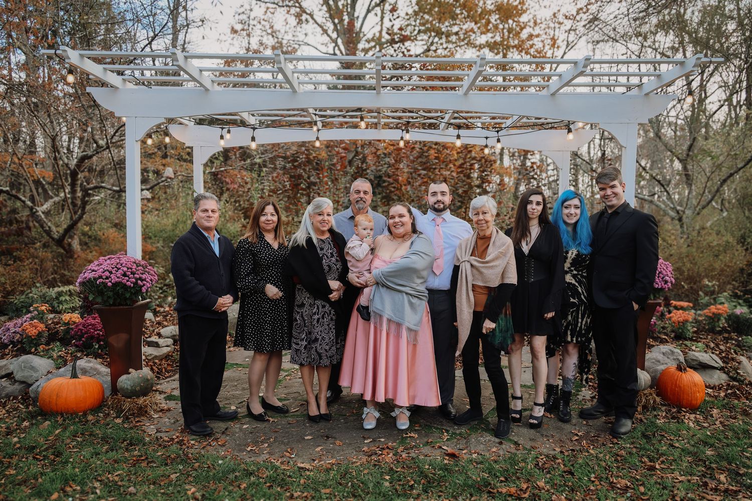 A group of people gather outdoors in autumn with pumpkins and fall foliage visible in the background.