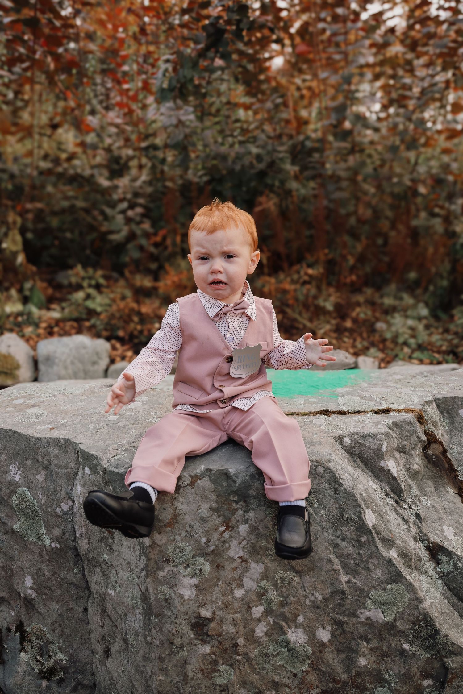 Child wearing pink coat sits on large gray rocks during autumn season with brown foliage in background.