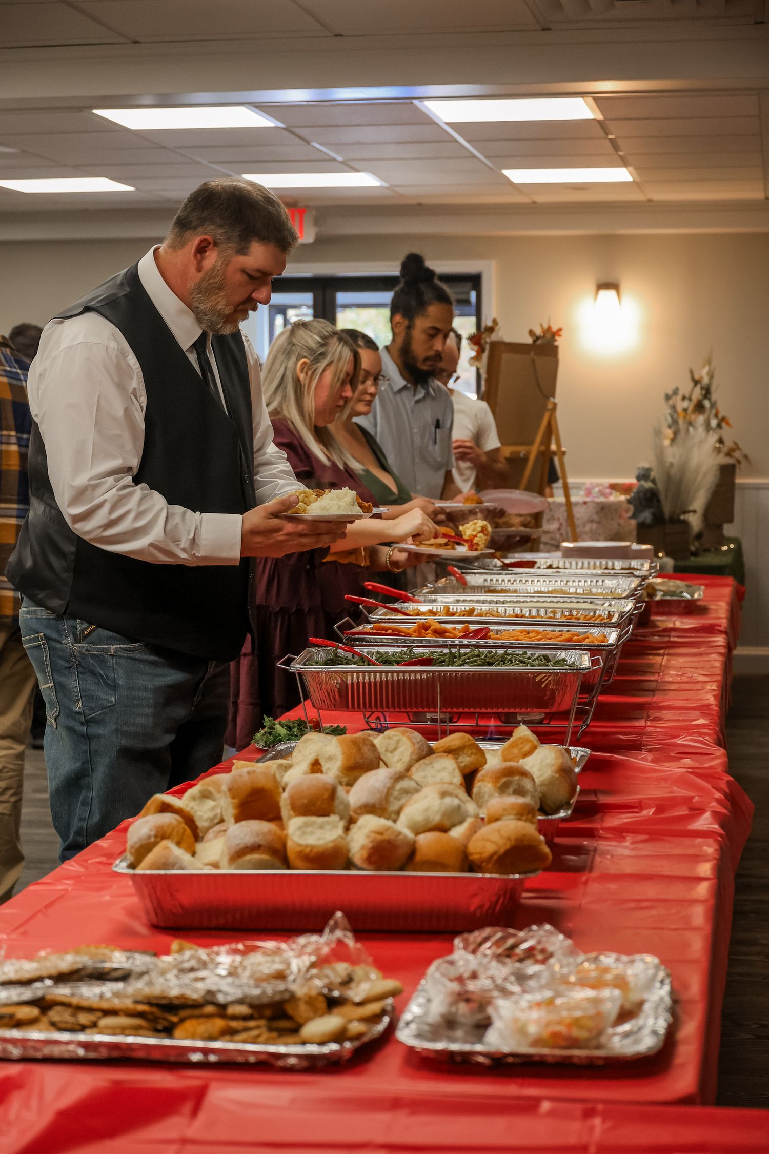 A buffet line with trays of food and rolls on a red tablecloth while people serve themselves at a catered event.