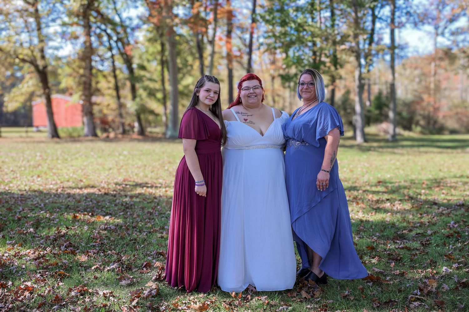 Four people wearing burgundy and blue dresses stand together in a fall outdoor setting with trees and leaves.