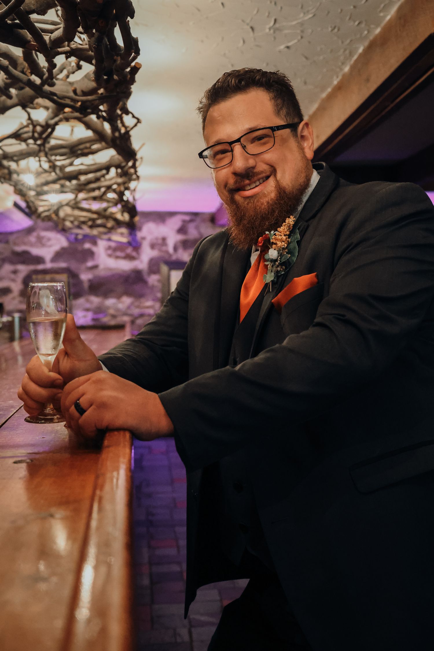 A wedding guest in a black suit with orange tie holds a champagne glass at a bar with purple ambient lighting.