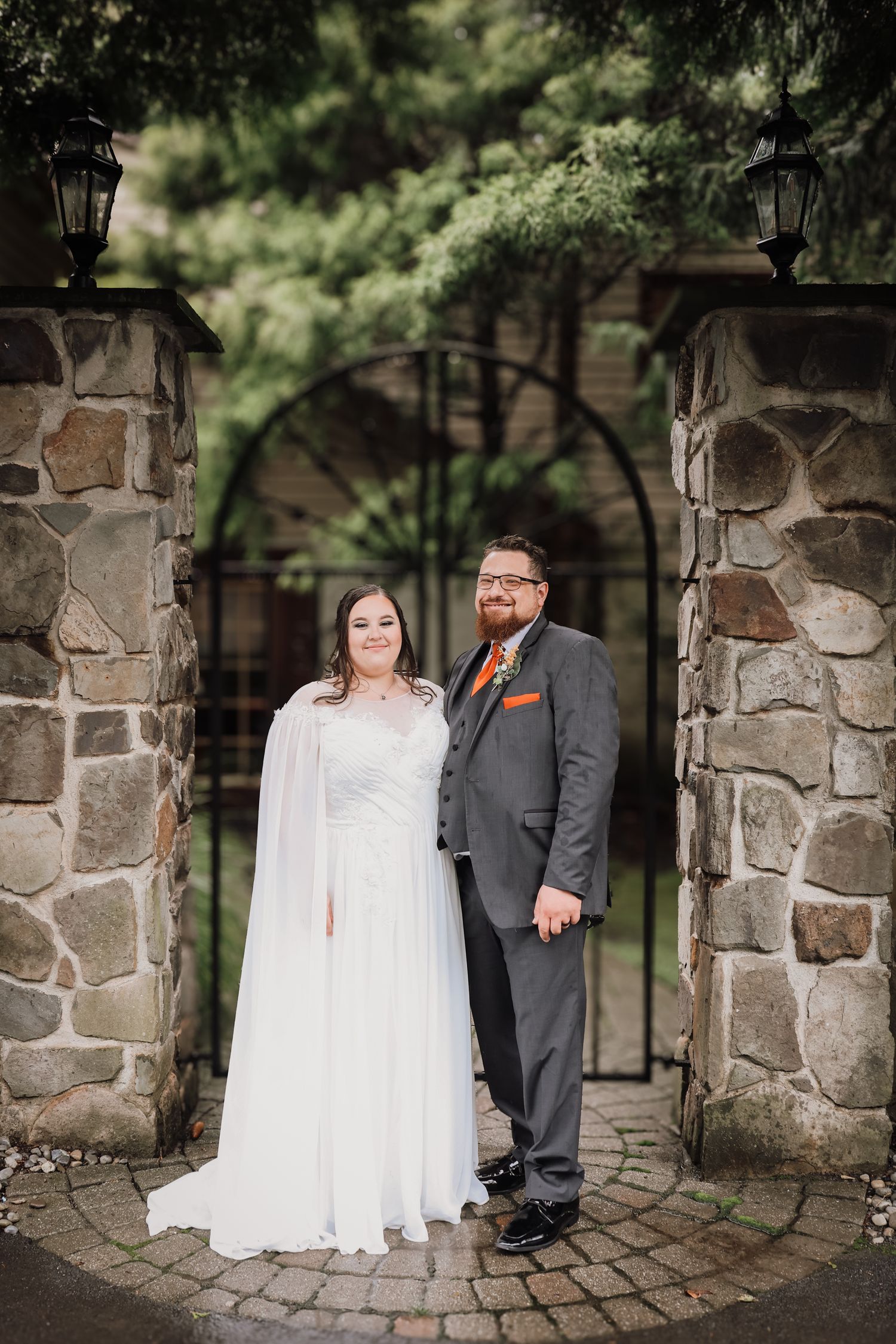 Couple in wedding attire poses against stone pillars and archways in a romantic outdoor garden setting.