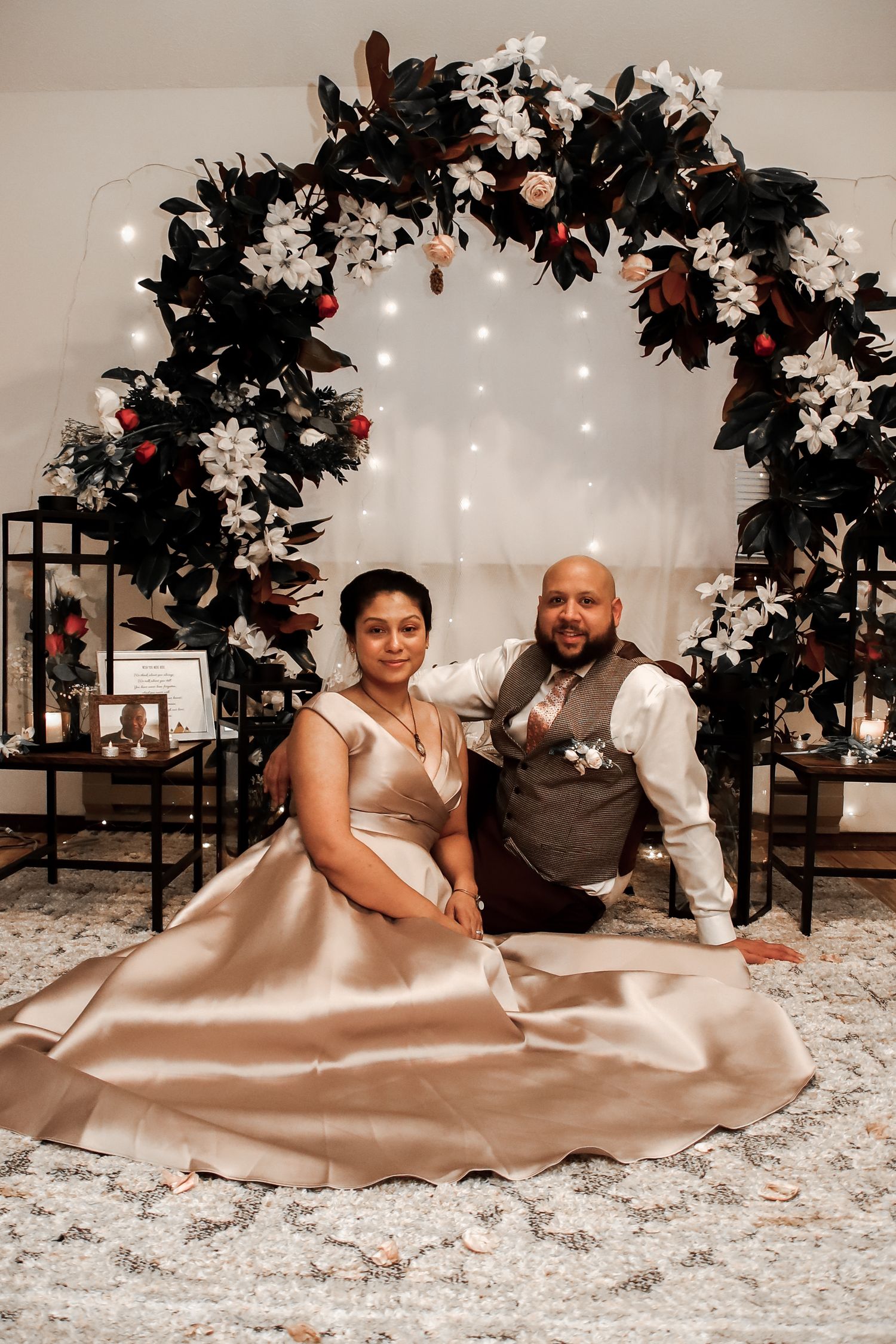 A couple in formal attire sits on a satin blanket surrounded by white snowflakes and dark foliage decorations.