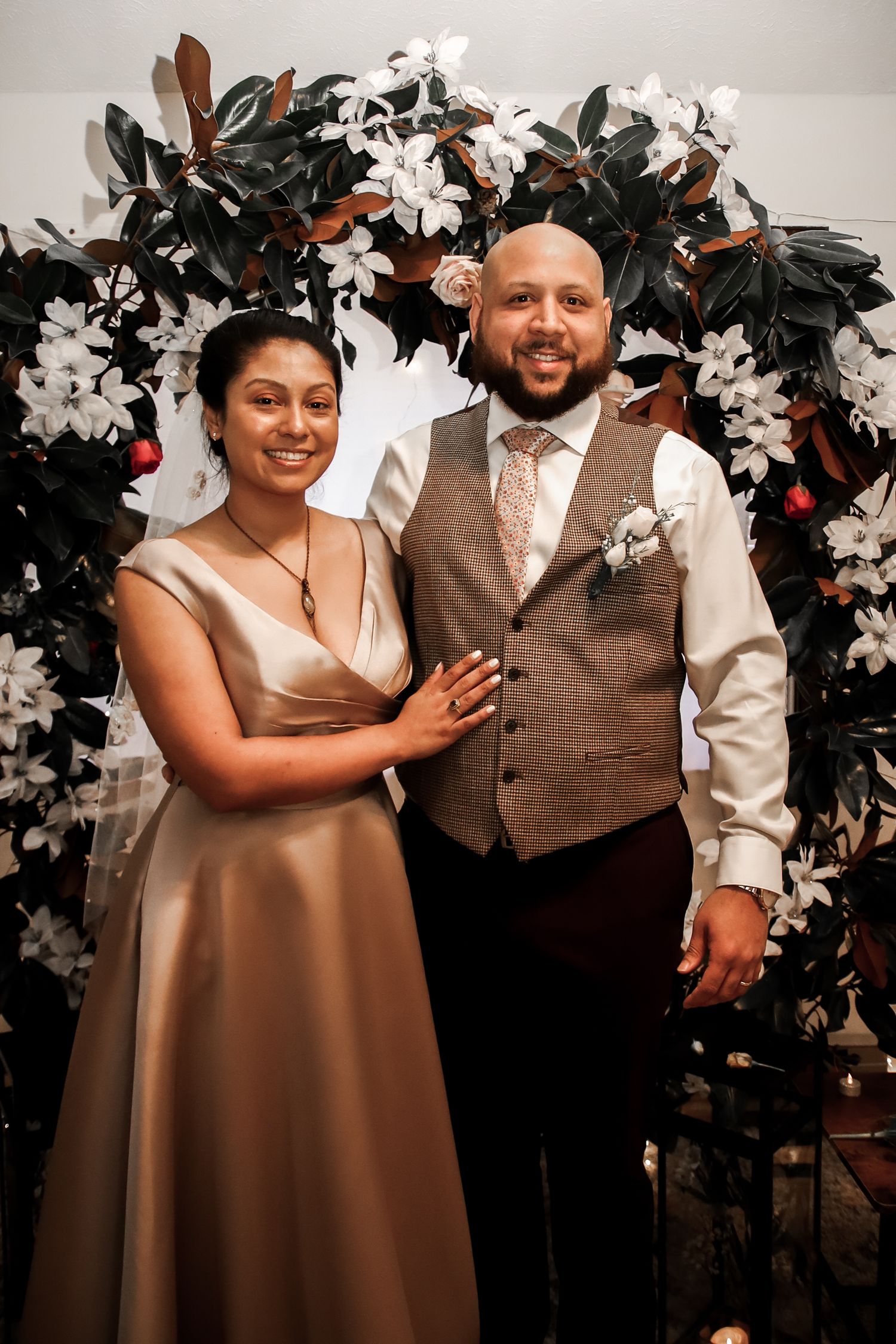 A couple poses in front of a floral arch with white flowers and dark leaves at an elegant wedding celebration.