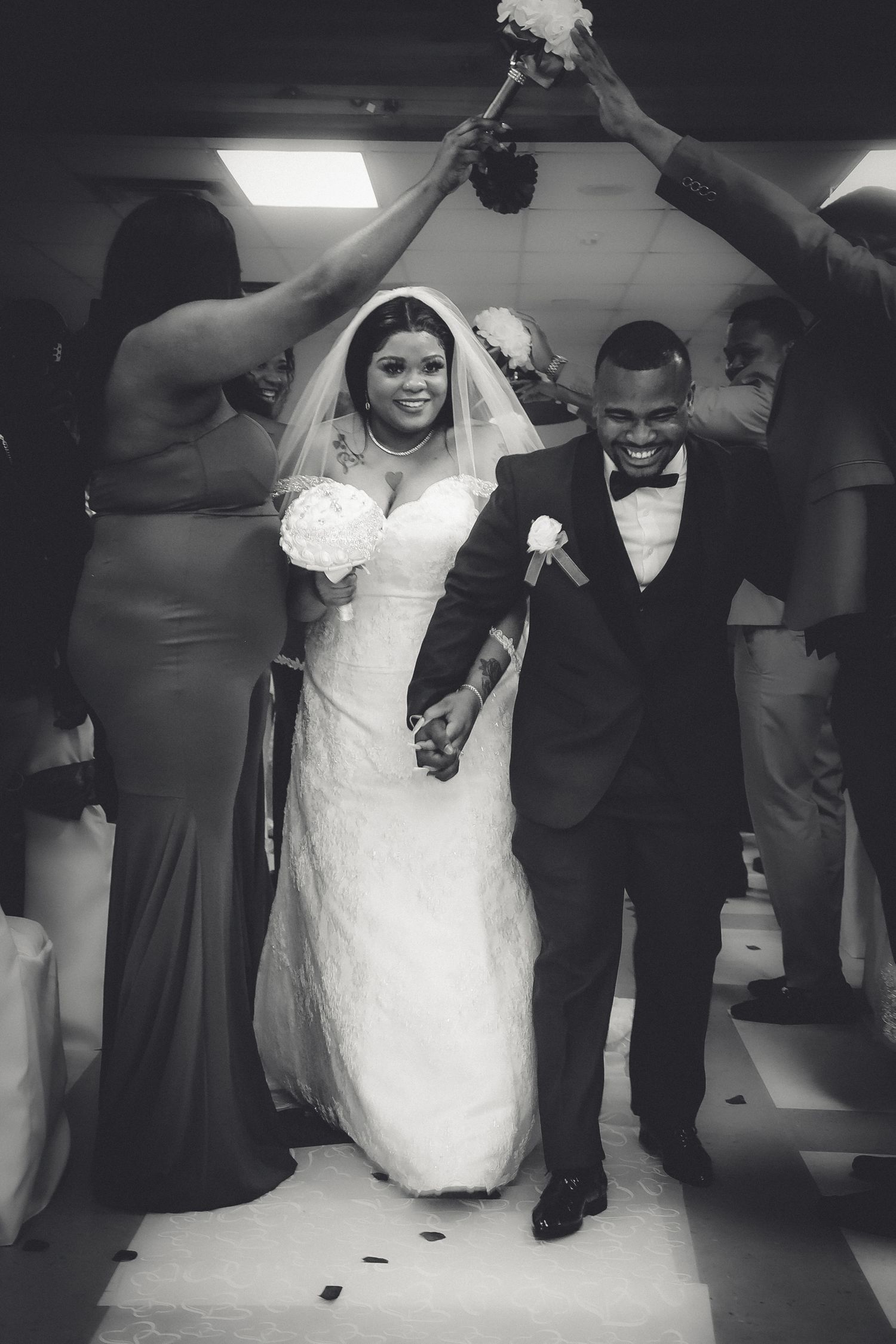 A joyful wedding couple walks down the aisle under raised hands during their ceremony celebration in black and white.