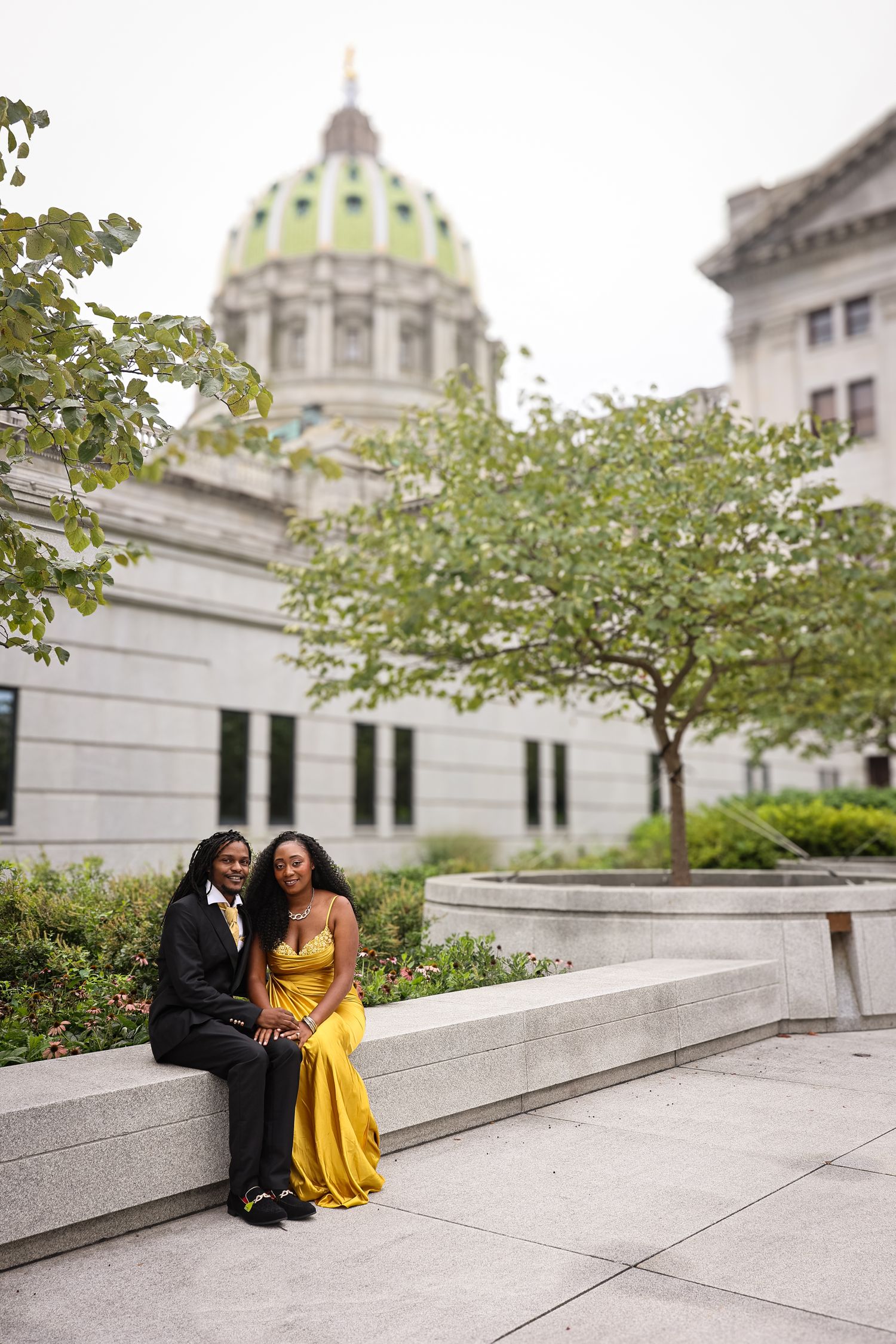 A couple sits together on stone steps outside a historic domed government building on an autumn day.