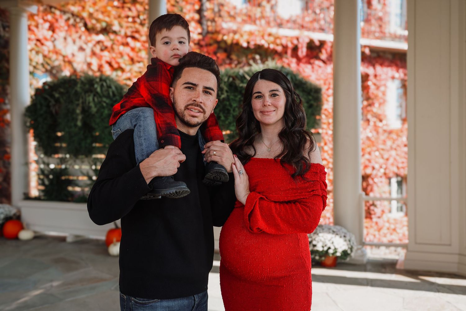 A couple in coordinating red and black outfits pose together on a porch with autumn foliage in the background.